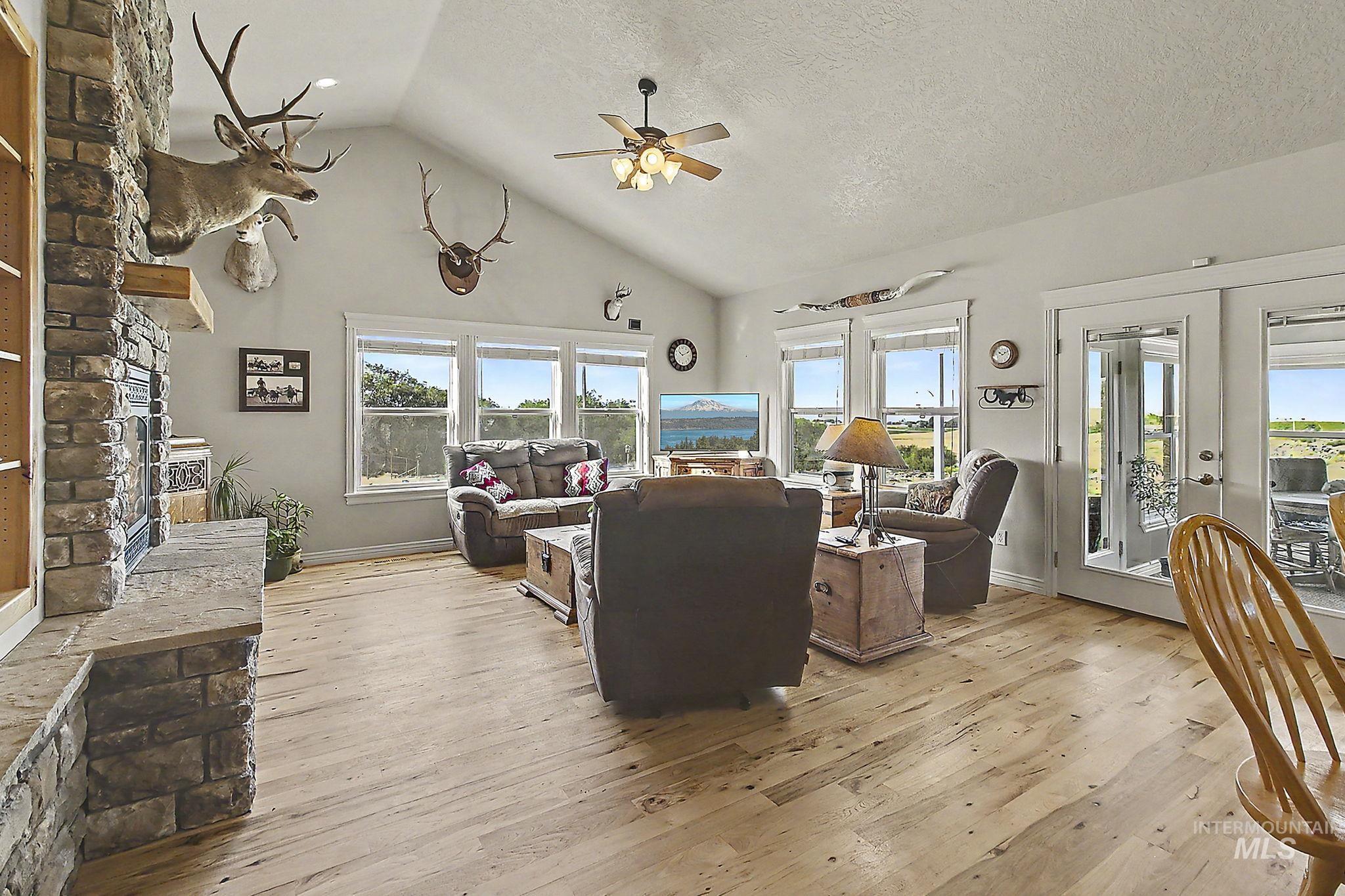 6900 West Charters Lane Melba, ID 83641 - Photo 8 of 49 Vaulted ceiling with a ceiling fan, light-toned wood-finish flooring, stone-clad fireplace with mantel, multiple windows, and exterior access doors