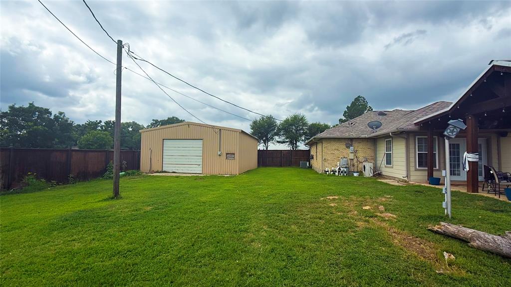 1356 Kerry Lane Azle, TX 76020 - Photo 25 of 27 Expansive backyard featuring a large metal utility building with a roll-up garage door, a covered patio area with exposed wood beams, and a partial privacy fence