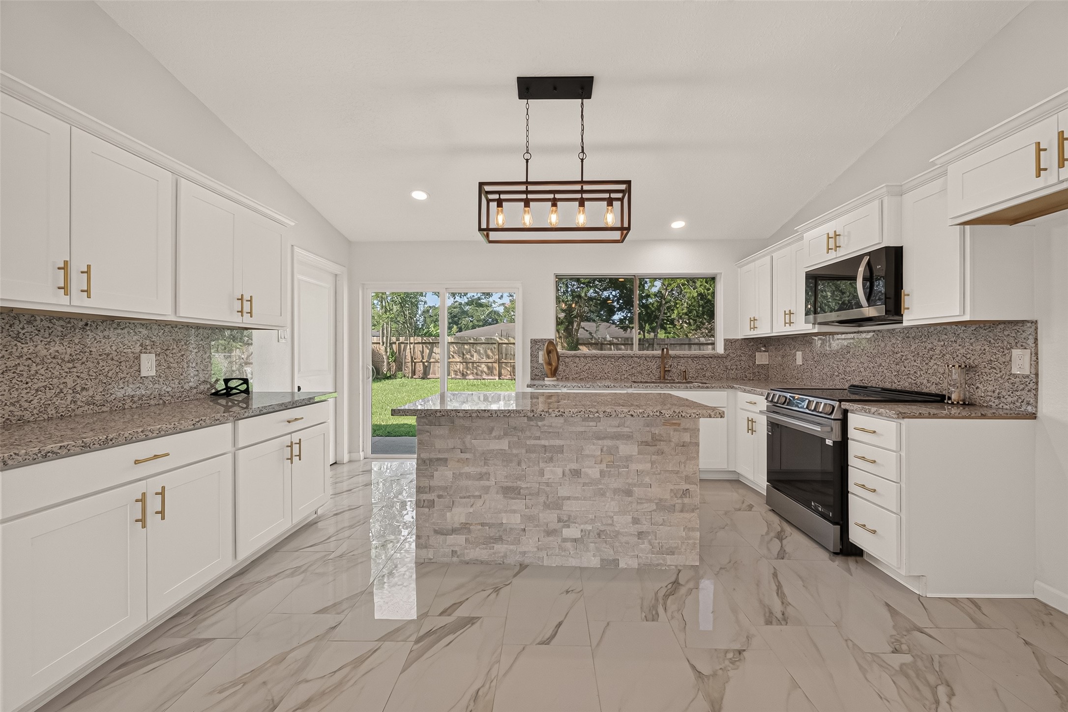 16619 Tibet Road Friendswood, TX 77546 - Photo 4 of 29 This modern kitchen features sleek white cabinetry with gold handles, granite countertops, and a central island with stone accents. It boasts stainless steel appliances, a stylish rectangular pendant light, and large windows allowing natural light to fill the space, complemented by polished marble-look tile flooring.