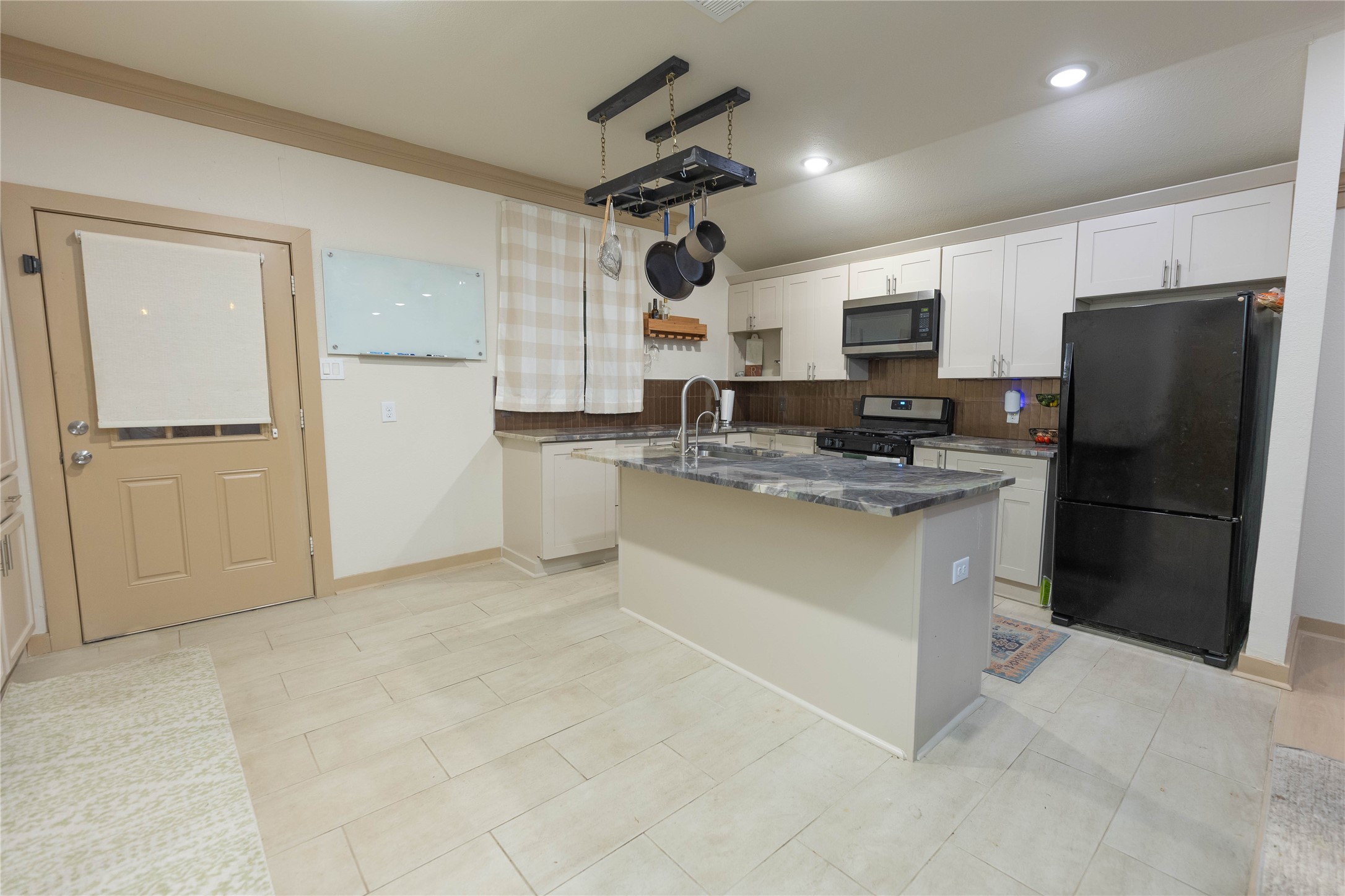 8007 Capitol Street Houston, TX 77012 - Photo 12 of 25 This modern kitchen features a central island with a marble countertop, stainless steel appliances, and ample white cabinetry. It includes a hanging pot rack, a black refrigerator, and a gas stove, offering a functional and stylish space for cooking.