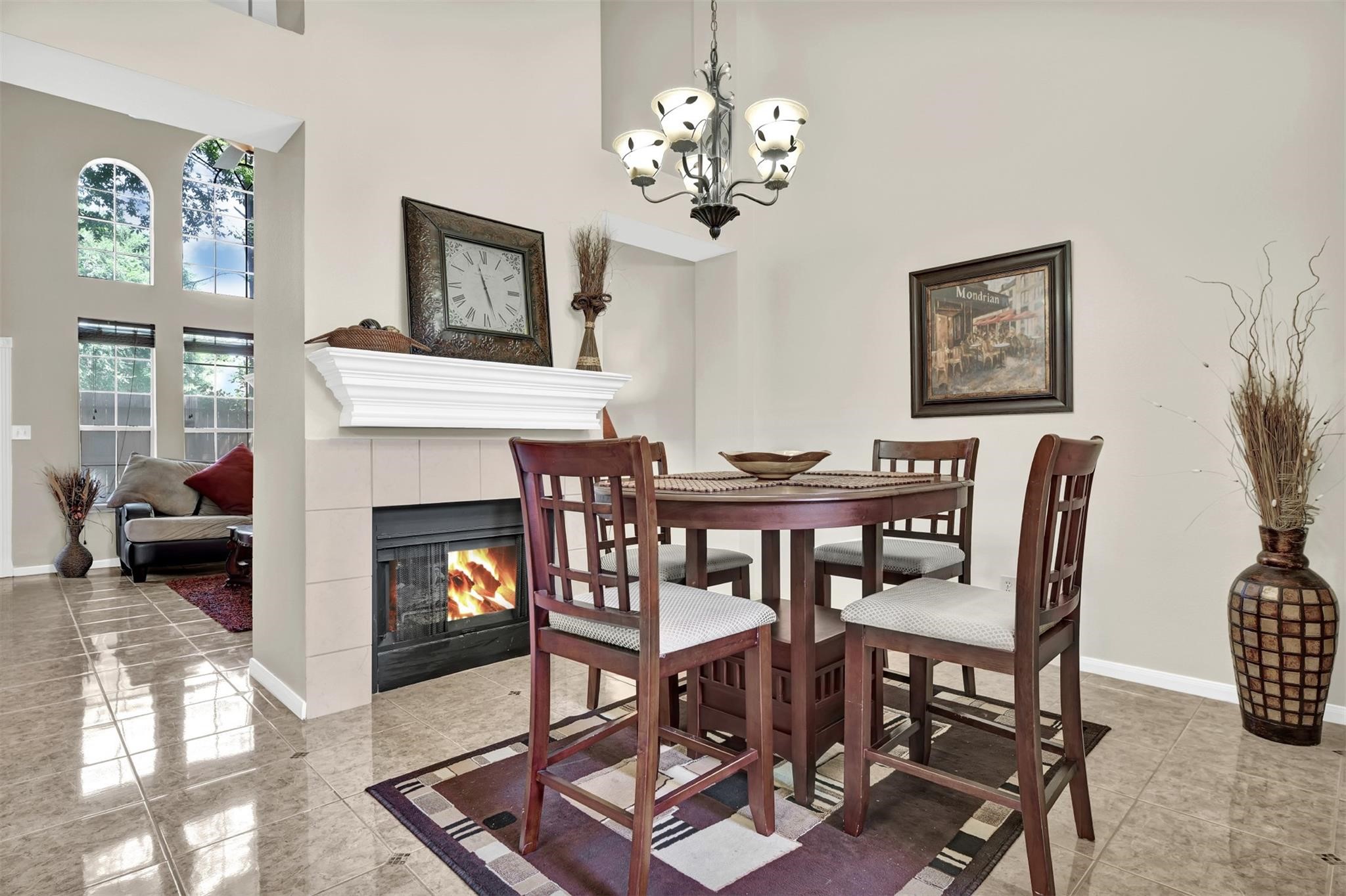14420 Walters Road, Unit 16 Houston, TX 77014 - Photo 8 of 27 This photo showcases a cozy dining area with a high ceiling, featuring a stylish chandelier and a round table with four chairs. The space is accentuated by a fireplace with a decorative mantel, tiled flooring, and an adjoining living area with large windows that bring in natural light.