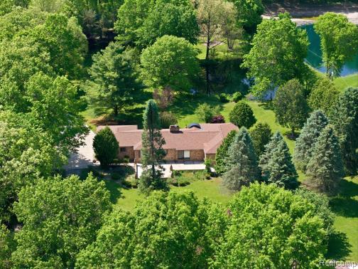 Ranch-style residence surrounded by mature trees, featuring a prominent brown shingle roof, brick exterior, and an expansive paved driveway