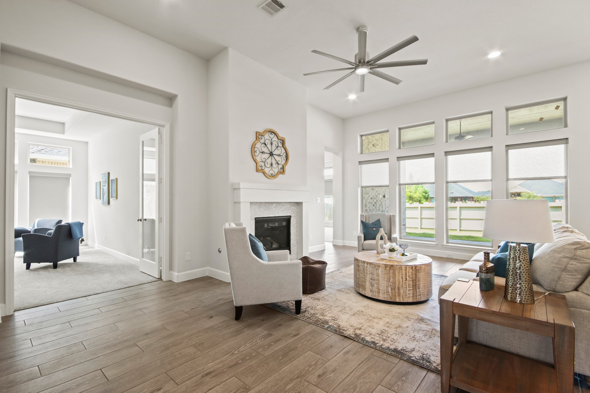 17406 Bayflower Conroe, TX 77385 - Photo 7 of 35 This inviting living room features a wall of windows for abundant natural light, a cozy fireplace, and a contemporary ceiling fan. The open layout leads to a bright adjacent room, perfect for flexible use. Stylish flooring and neutral tones create a warm, modern ambiance.