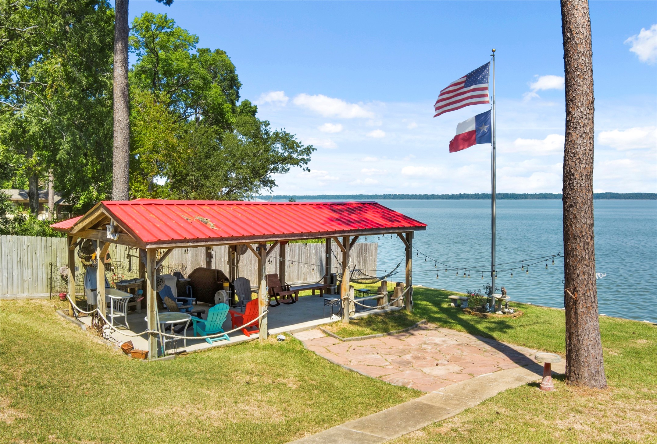 200 Trott Lane Point Blank, TX 77364 - Photo 40 of 50 Lakeside relaxation at its best! This covered pavilion with a vibrant red metal roof offers a perfect spot for outdoor entertaining, complete with ample seating, string lights, and unbeatable water views. Enjoy gentle breezes off the lake while you unwind under the shade or gather with family and friends for sunset evenings by the shore.