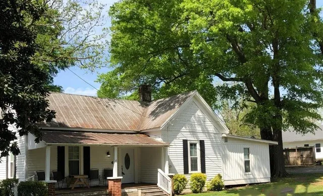 a view of a white house next to a yard with plants and large trees