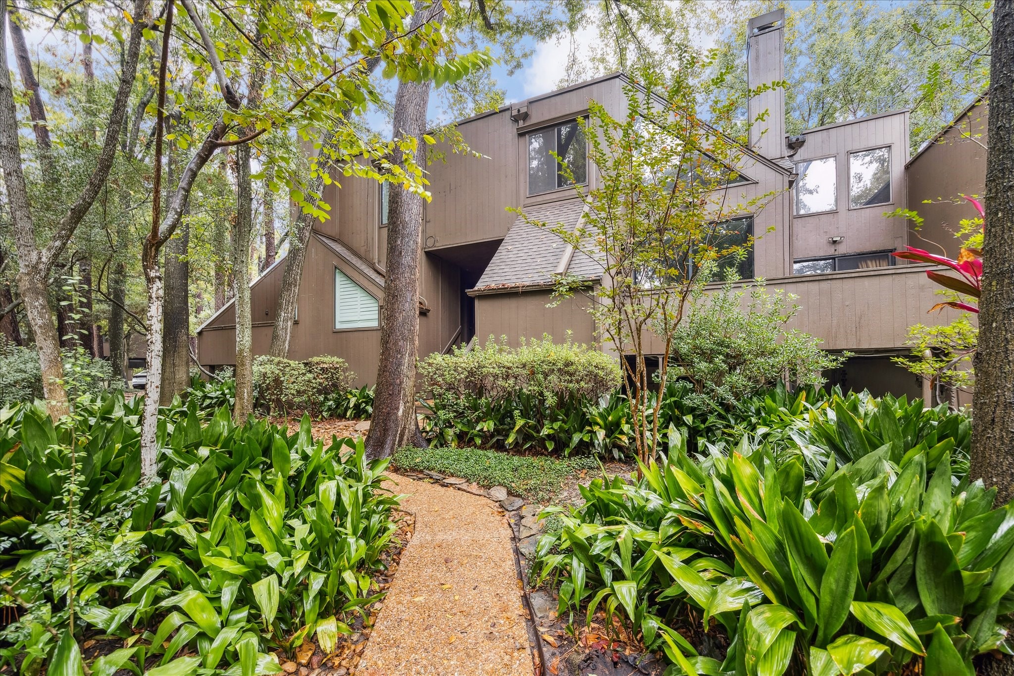 Charming wooded walkway leads to this contemporary townhome, surrounded by lush landscaping and mature trees. The inviting brick path is framed by vibrant greenery, creating a serene and private entrance