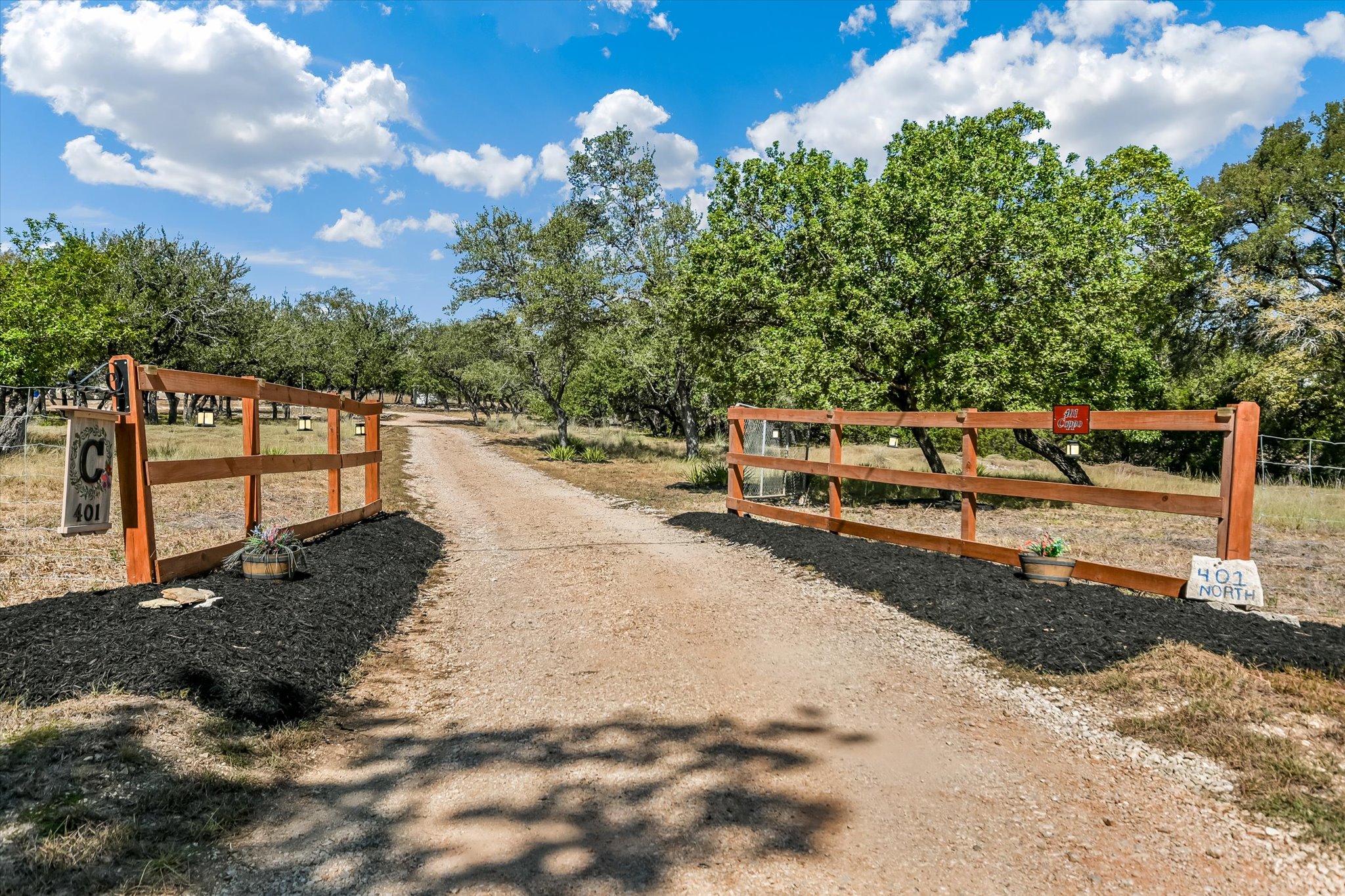 401 North Rainbow Ranch Road Wimberley, TX 78676 - Photo 1 of 39 A rustic wood-fenced entrance opens to a long gravel driveway shaded by mature oak trees.
