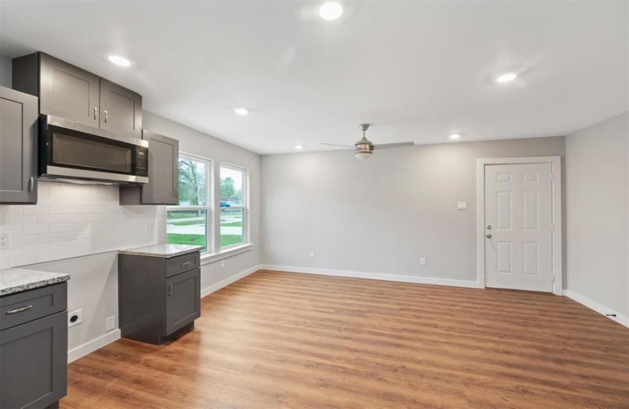 8120 Laura Koppe Road, Unit B Houston, TX 77028 - Photo 6 of 25 This photo showcases a modern open-concept living area with hardwood floors and a sleek kitchen featuring dark cabinetry, a built-in microwave, and light countertops. Large windows provide natural light, and a ceiling fan adds comfort.