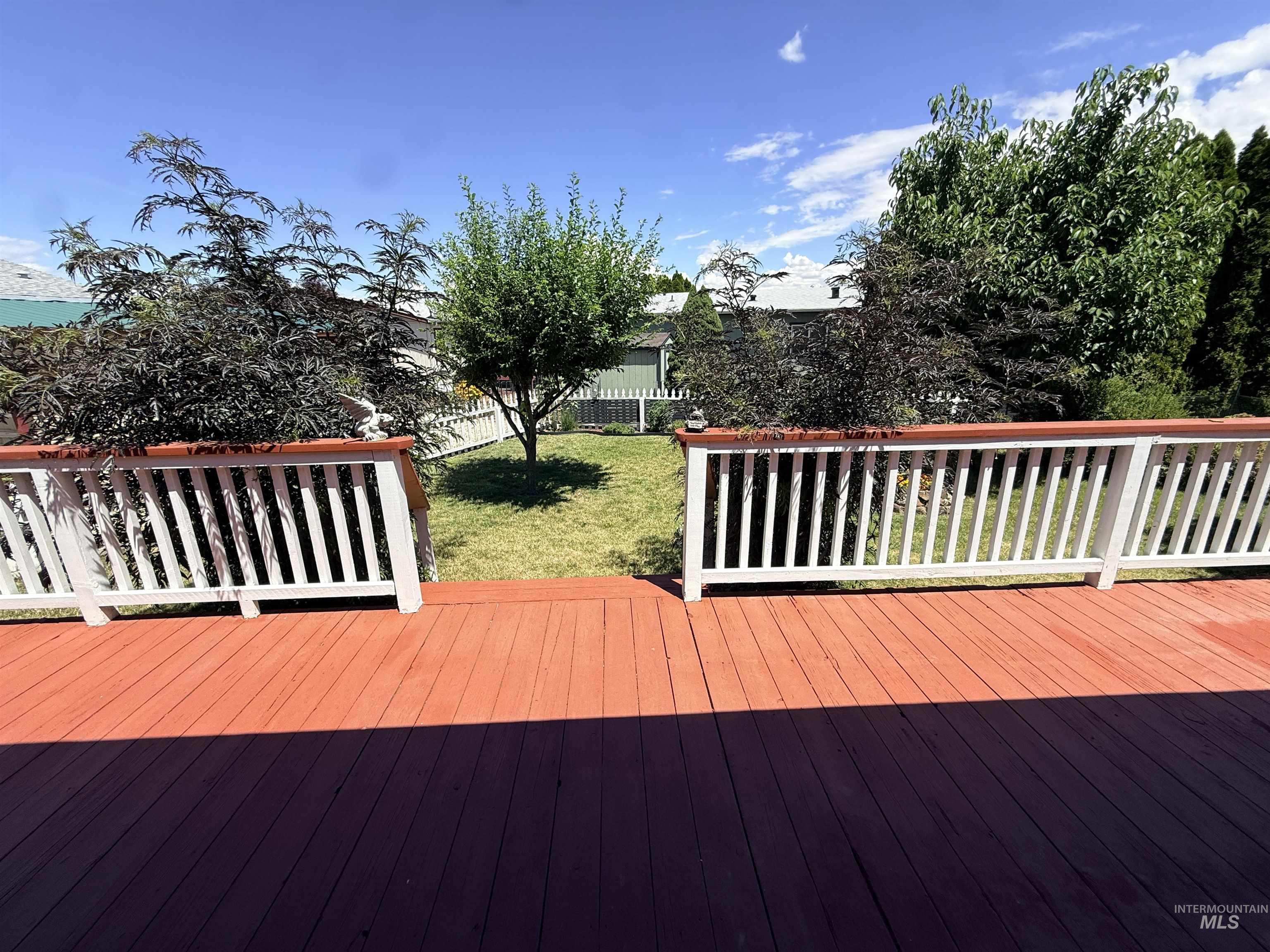 2750 Alden Road, Unit 12 12 Fruitland, ID 83619 - Photo 37 of 38 Expansive backyard featuring a vibrant red-stained deck with white railings, a manicured lawn, established trees, and a white picket fence