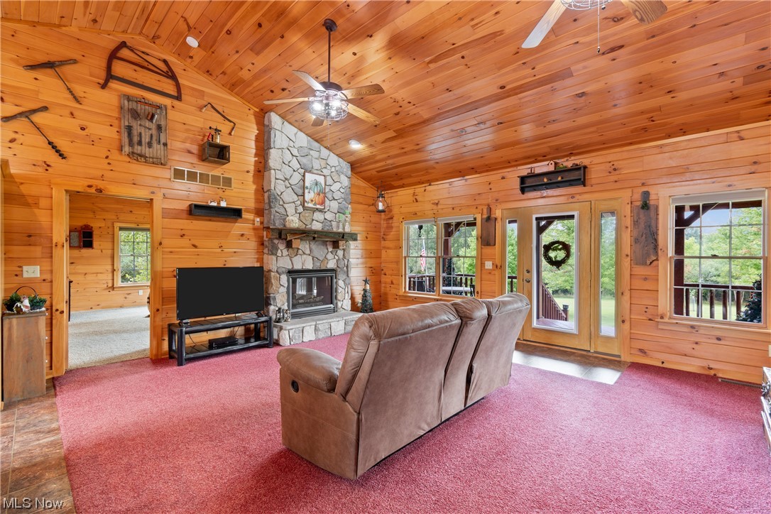 Carpeted living room featuring wood ceiling, vaulted ceiling, plenty of natural light, ceiling fan, a stone fireplace, and wooden walls