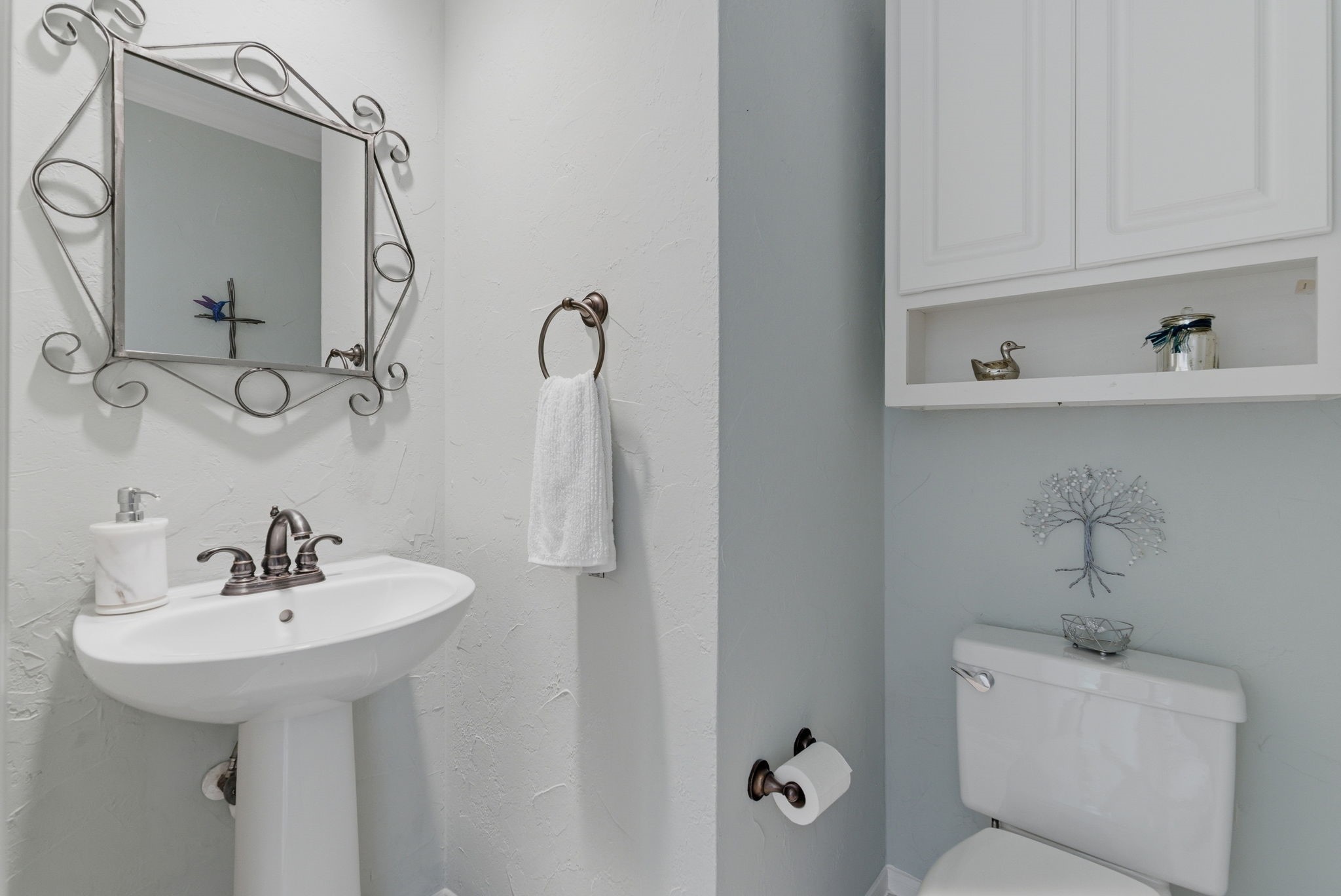 11 Dovewing Place Spring, TX 77382 - Photo 25 of 32 This is a small, elegant bathroom with a pedestal sink, ornate mirror, and soft lighting. It features a towel ring, a toilet with overhead cabinet storage, and simple decorative elements.