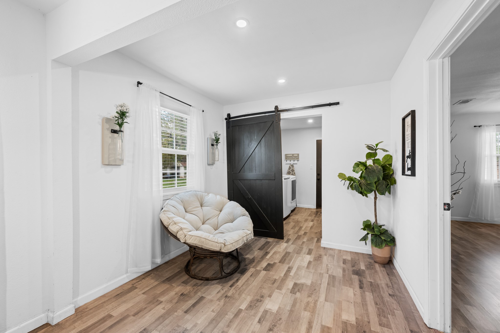 729 Kyle Angleton, TX 77515 - Photo 18 of 36 This bright, inviting space features hardwood-look floors, a cozy chair by a window with sheer curtains, a stylish barn door, and tasteful decor including plants, creating a warm and welcoming atmosphere.