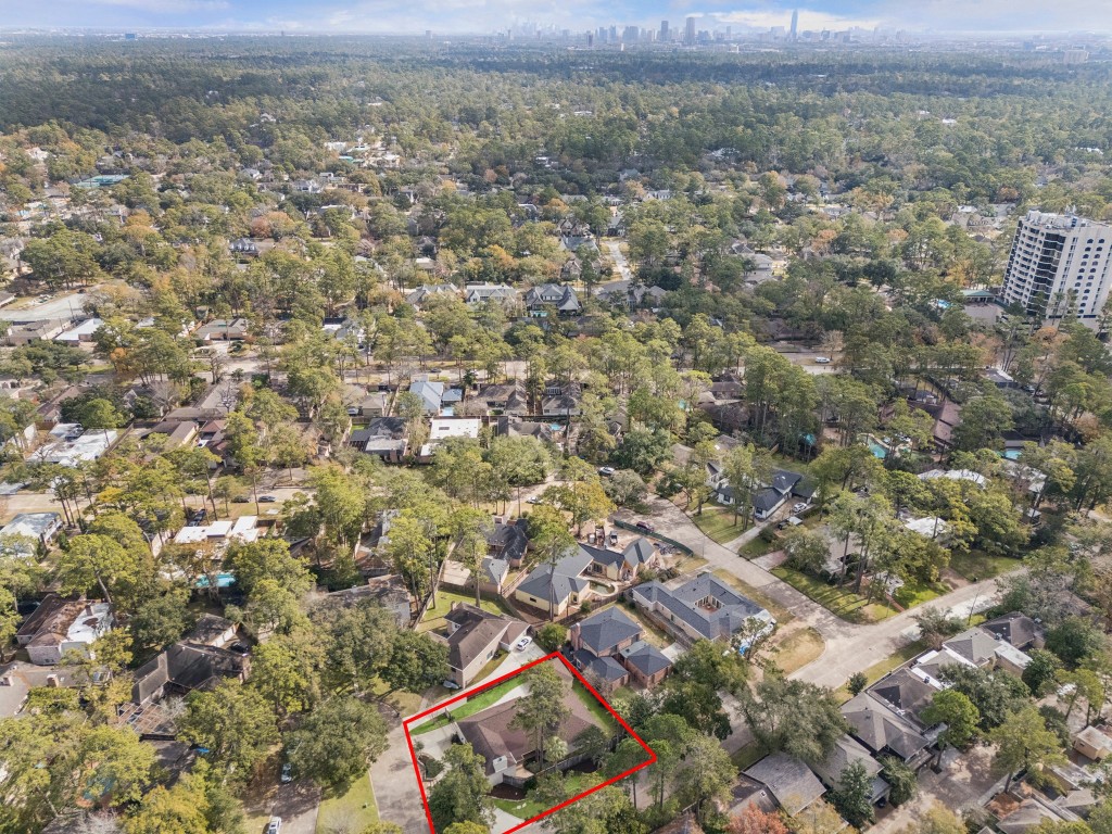 242 Vanderpool Lane Houston, TX 77024 - Photo 38 of 45 This aerial photo showcases a property within a lush, tree-filled neighborhood, marked by a red outline. The area appears residential, with well-spaced houses and a mix of modern and traditional architecture. The view extends to a distant city skyline, indicating proximity to urban amenities while maintaining a serene suburban feel.