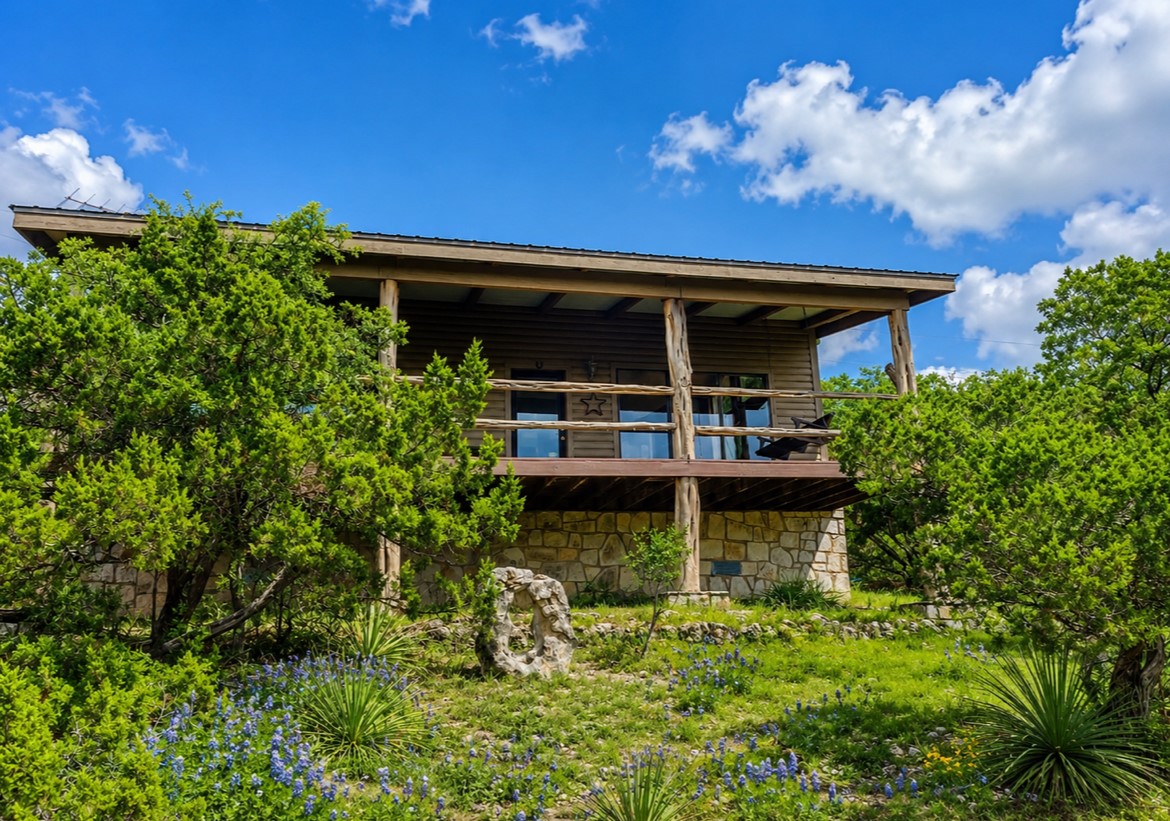 919 Little Ranches Road Wimberley, TX 78676 - Photo 2 of 13 A charming cabin, nestled amongst flourishing greenery, features a welcoming covered balcony and stone foundation.