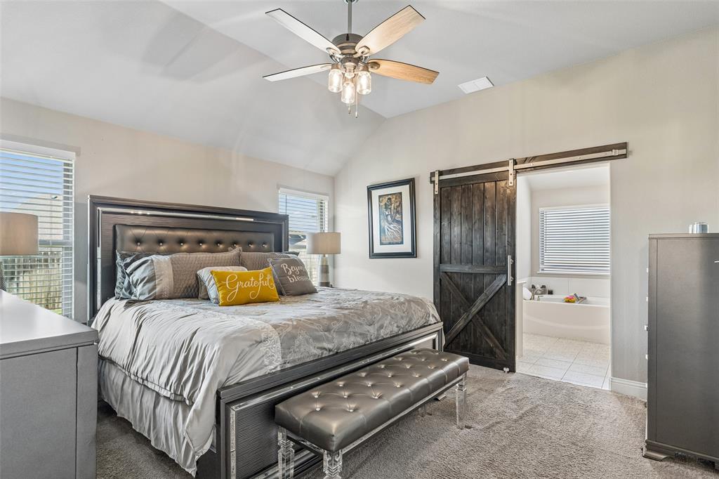 108 Maverick Lane Pilot Point, TX 76258 - Photo 16 of 27 Spacious bedroom featuring vaulted ceilings, a ceiling fan with integrated lighting, two windows with blinds, plush carpet flooring, and a dark wood-finish barn door opening to a bathroom with a soaking tub