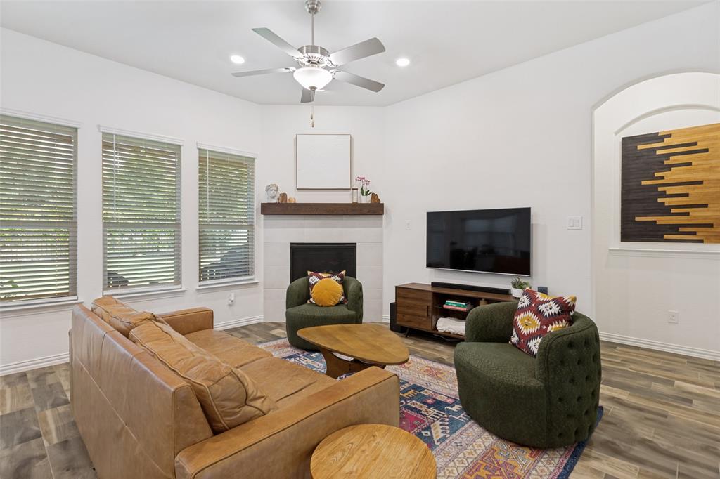 5556 Vaquero Road Fort Worth, TX 76126 - Photo 8 of 25 Living area featuring a fireplace with a wood mantle, three large windows with blinds, a ceiling fan with integrated lighting, and wood-finish flooring