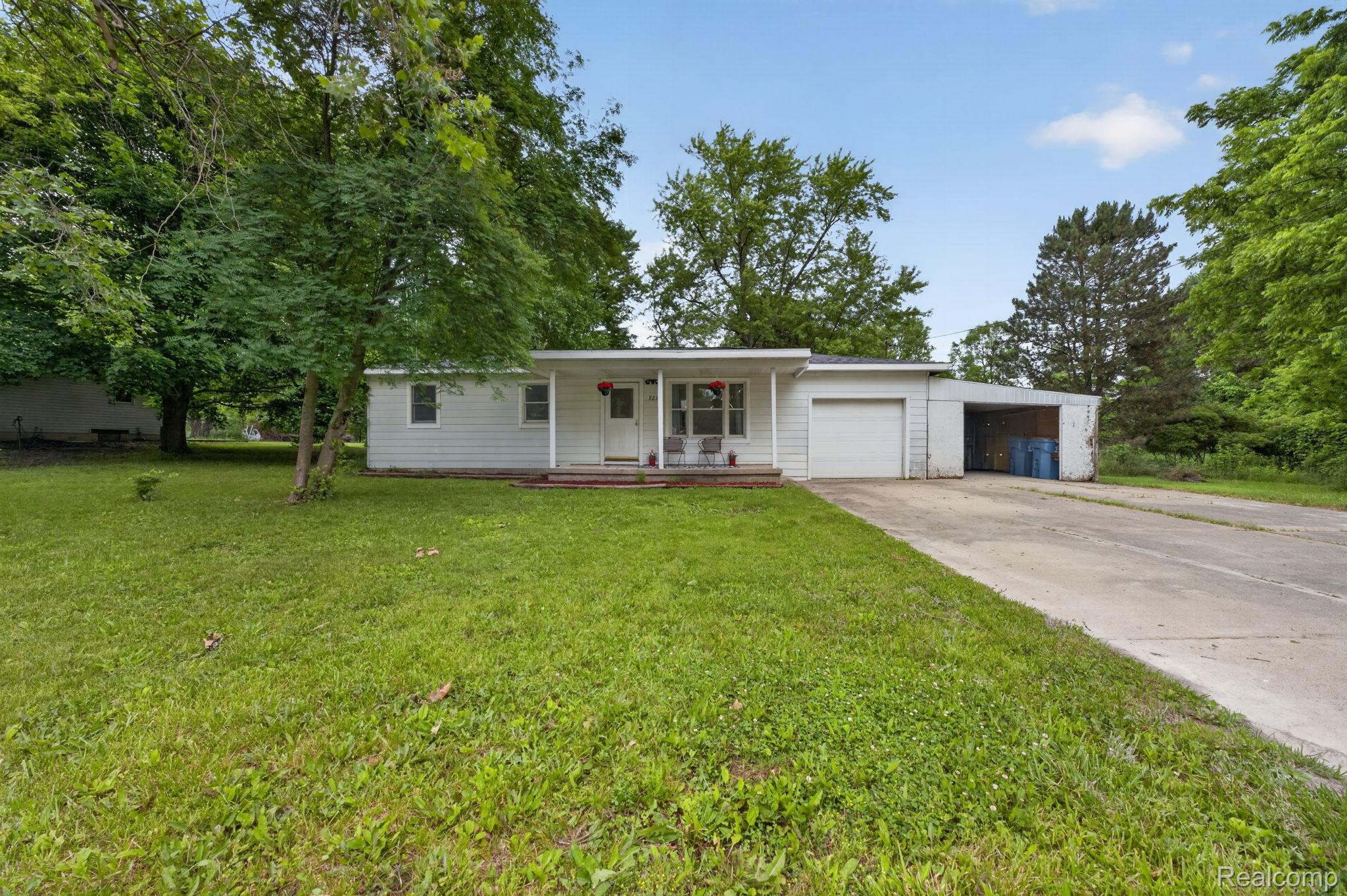9288 Hilda Lane Flushing, MI 48433 - Photo 6 of 50 Single-story white siding residence featuring a covered front porch, an attached garage, a secondary carport structure, a concrete driveway, and mature landscaping