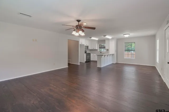 an empty room with wooden floor ceiling fan and kitchen view