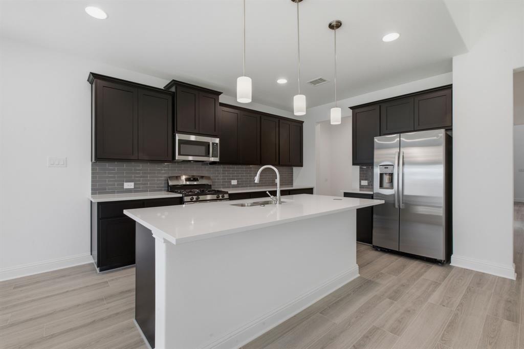 2911 Hanscom Street Forney, TX 75126 - Photo 22 of 40 Kitchen featuring dark wood cabinetry, stainless steel appliances, a white island with an undermount sink, a gray subway tile backsplash, and light wood-finish flooring