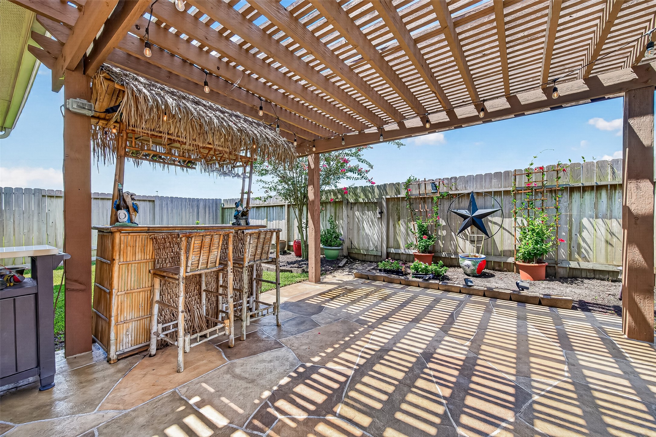 4910 Culver Lane Rosenberg, TX 77471 - Photo 5 of 50 This outdoor space features a wooden pergola with a Tiki bar (excluded) and stools (excluded,) perfect for entertaining. The area is accented with a stone patio floor, colorful garden beds, and decorative elements, creating a relaxing backyard retreat (plants in pots and pots are excluded.)