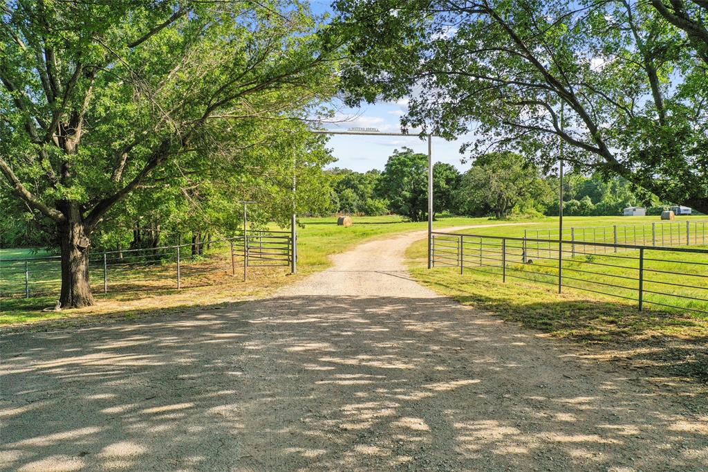 3030 Wager Road Flower Mound, TX 75028 - Photo 13 of 40 Gravel driveway entry framed by mature trees, featuring a metal gate and fencing bordering expansive green pastures