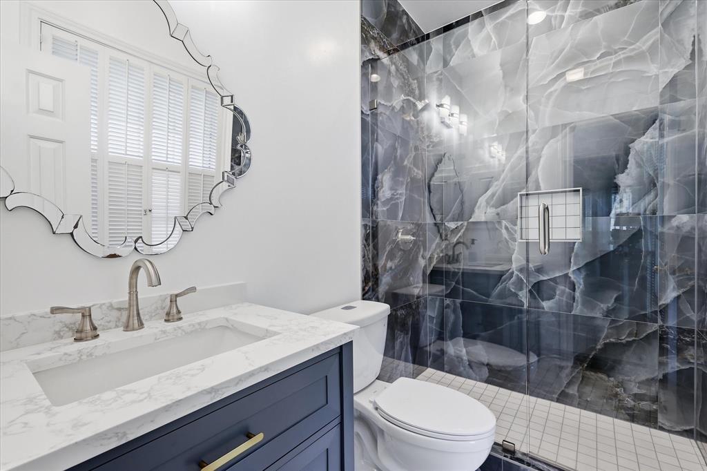 6955 Hawk Road Flower Mound, TX 75022 - Photo 28 of 40 Modern bathroom featuring a glass-enclosed shower with large format marble-look tile, a white vanity countertop, a deep blue vanity cabinet with gold-tone hardware, and a decorative beveled mirror