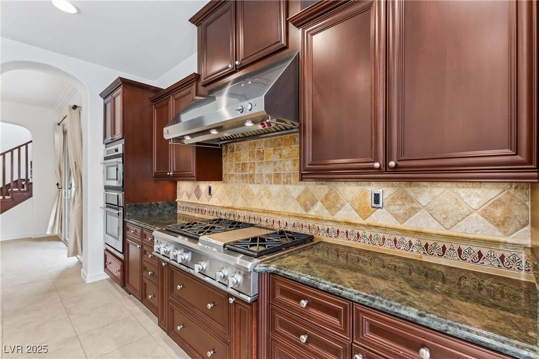 58 Contrada Fiore Drive Henderson, NV 89011 - Photo 13 of 56 Kitchen featuring arched walkways, under cabinet range hood, dark stone countertops, appliances with stainless steel finishes, and ornamental molding