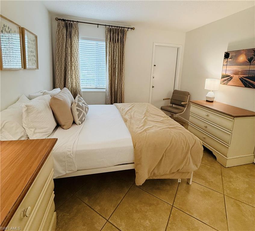 3250 10th Street North, Unit C8 Naples, FL 34103 - Photo 7 of 24 Bedroom featuring light-toned tile flooring, a window with blinds and patterned drapes, a white paneled door, and neutral-toned wall paint