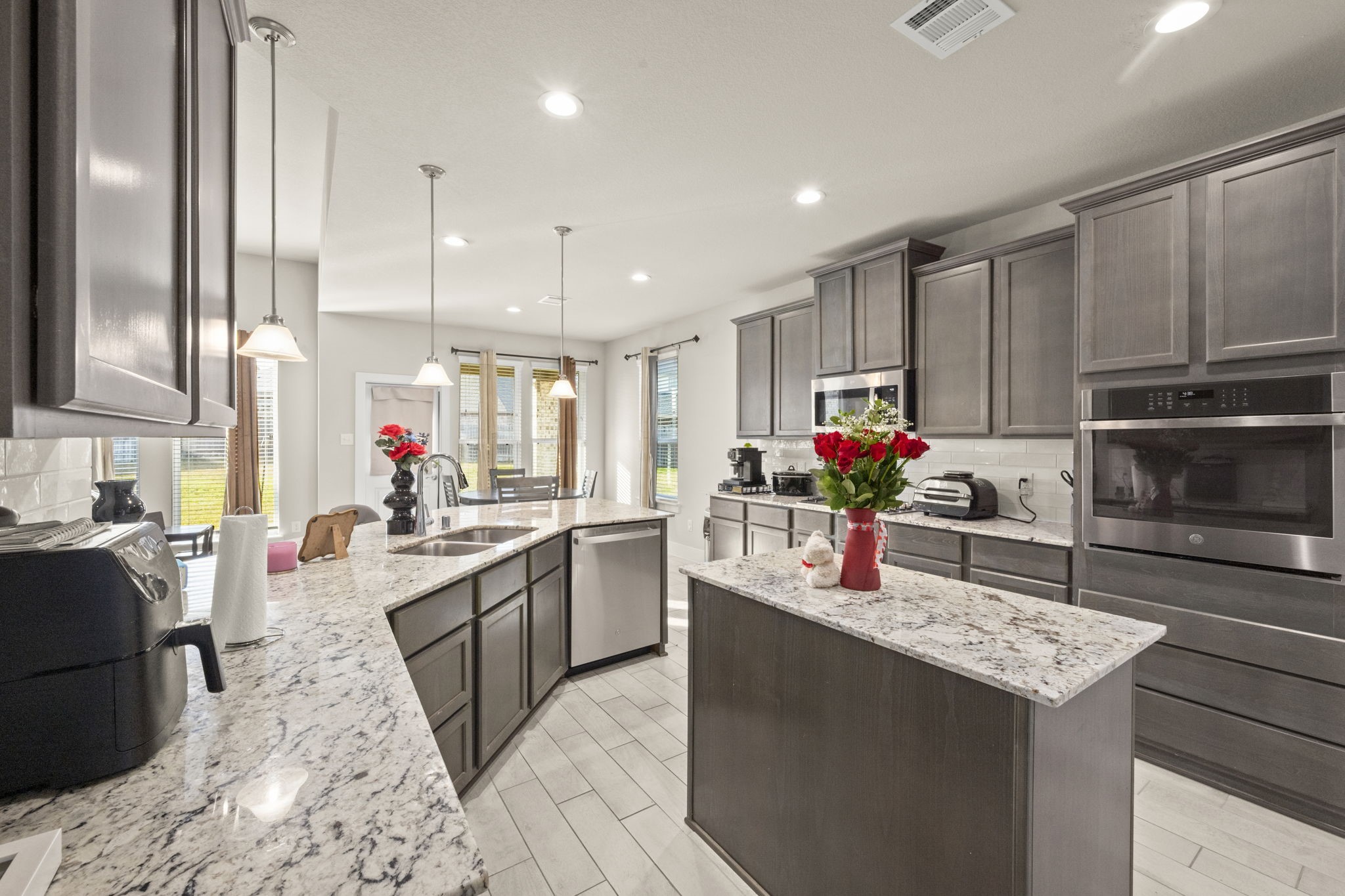 24715 Native Forest Spring, TX 77373 - Photo 11 of 37 This spacious, modern kitchen features sleek dark cabinetry, granite countertops, and stainless steel appliances. It boasts a large island with seating, abundant natural light from multiple windows, and stylish pendant lighting, creating an inviting cooking and dining space.