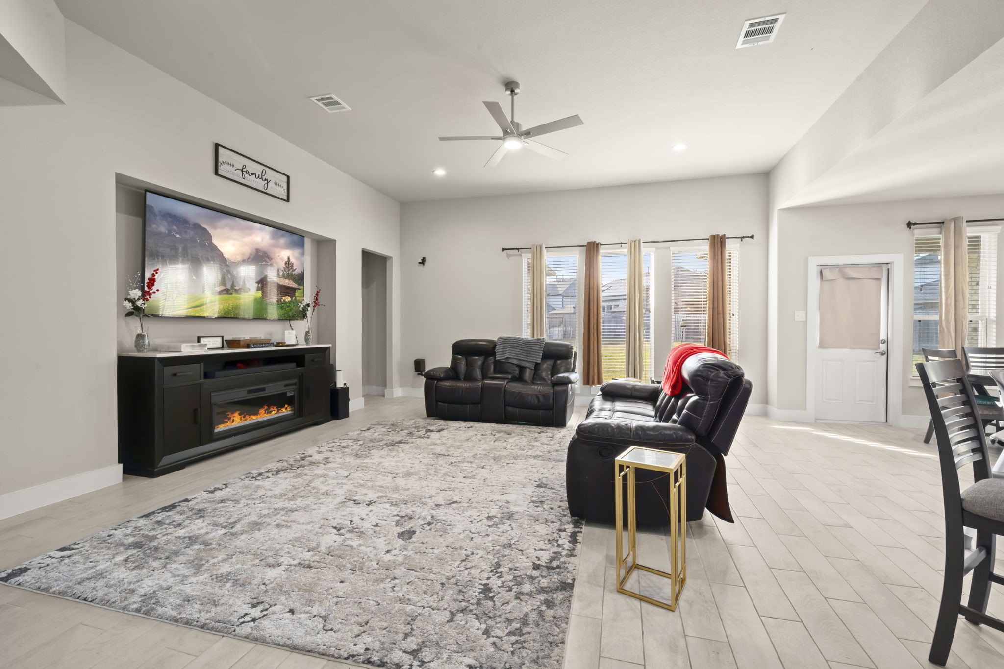24715 Native Forest Spring, TX 77373 - Photo 15 of 37 This spacious living room features a modern design with neutral tones, high ceilings, and ample natural light. It includes a large wall-mounted TV, an electric fireplace, and comfortable leather seating. The room opens to a dining area and has access to the backyard through glass doors, offering a perfect blend of style and functionality for family gatherings.