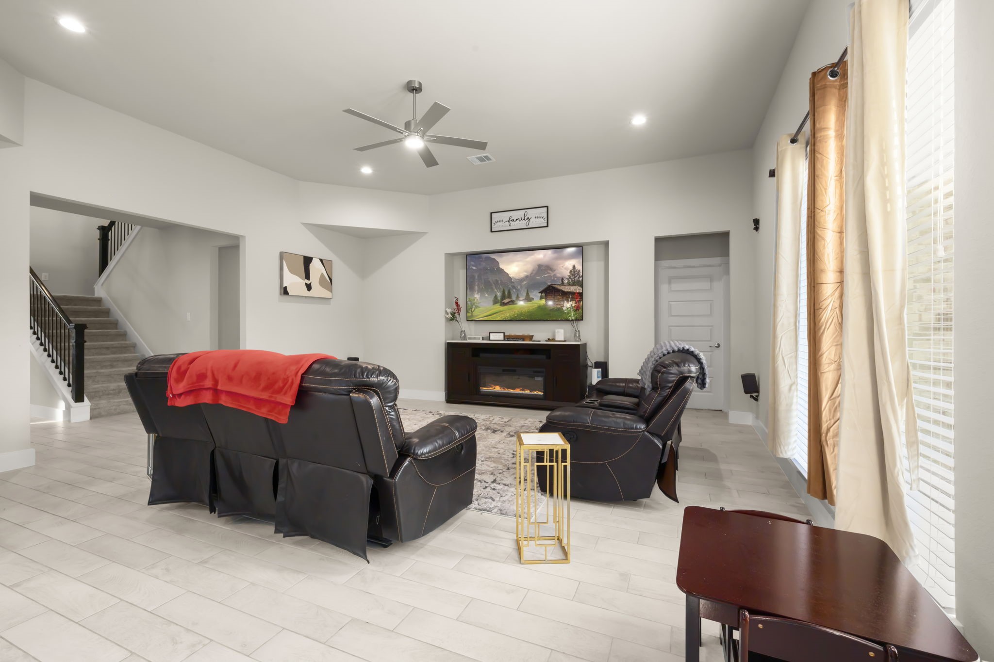 24715 Native Forest Spring, TX 77373 - Photo 16 of 37 This spacious living room features modern tile flooring, ample natural light from large windows, and a neutral color palette. It includes a cozy seating area, a flat-screen TV above a fireplace, and access to a carpeted staircase leading upstairs.