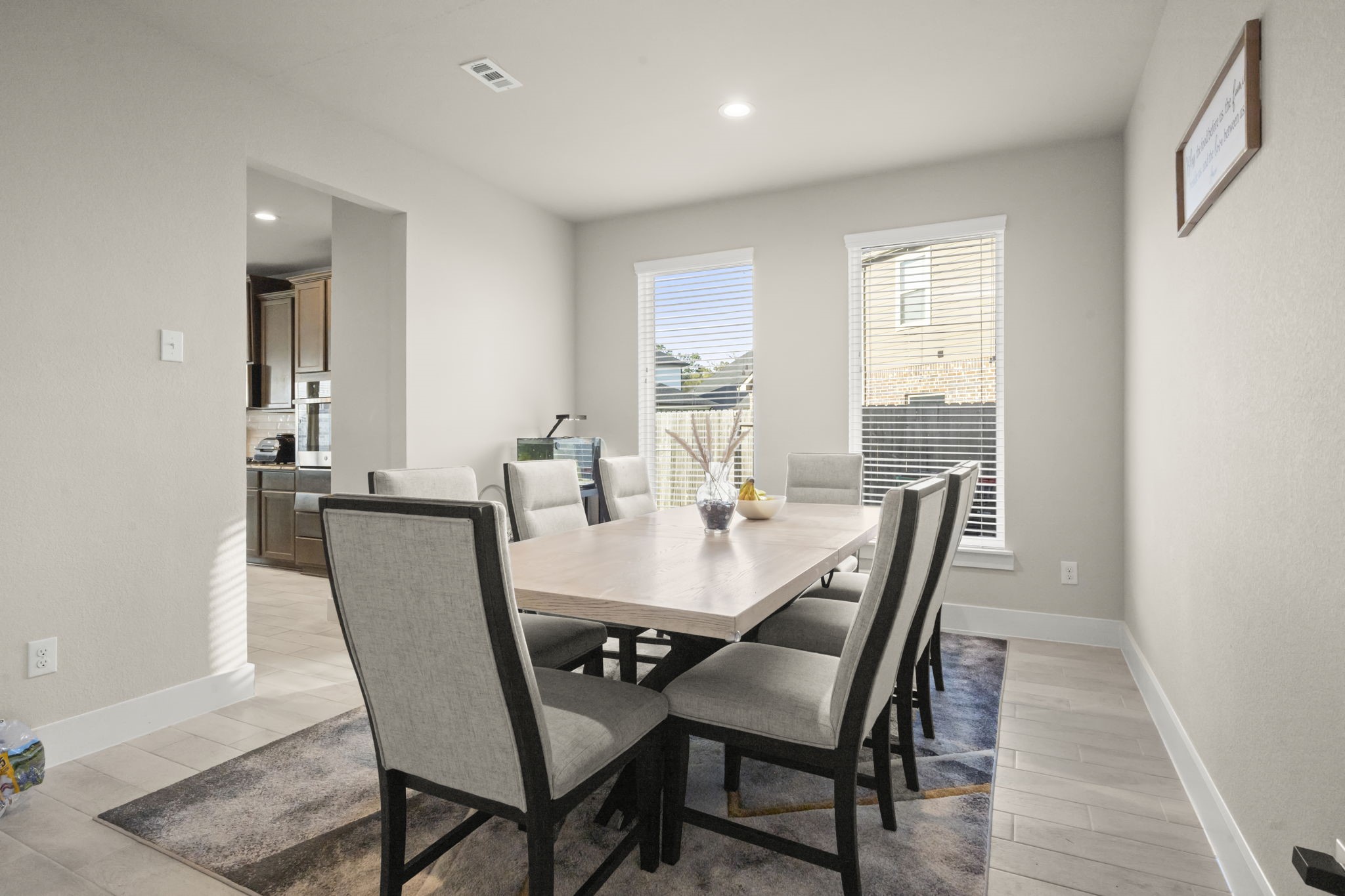 24715 Native Forest Spring, TX 77373 - Photo 7 of 37 This photo shows a bright, modern dining room with a large table and eight chairs. The room features light wood flooring, neutral walls, and two windows that let in natural light. There's an open doorway leading to the kitchen, adding to the spacious feel.