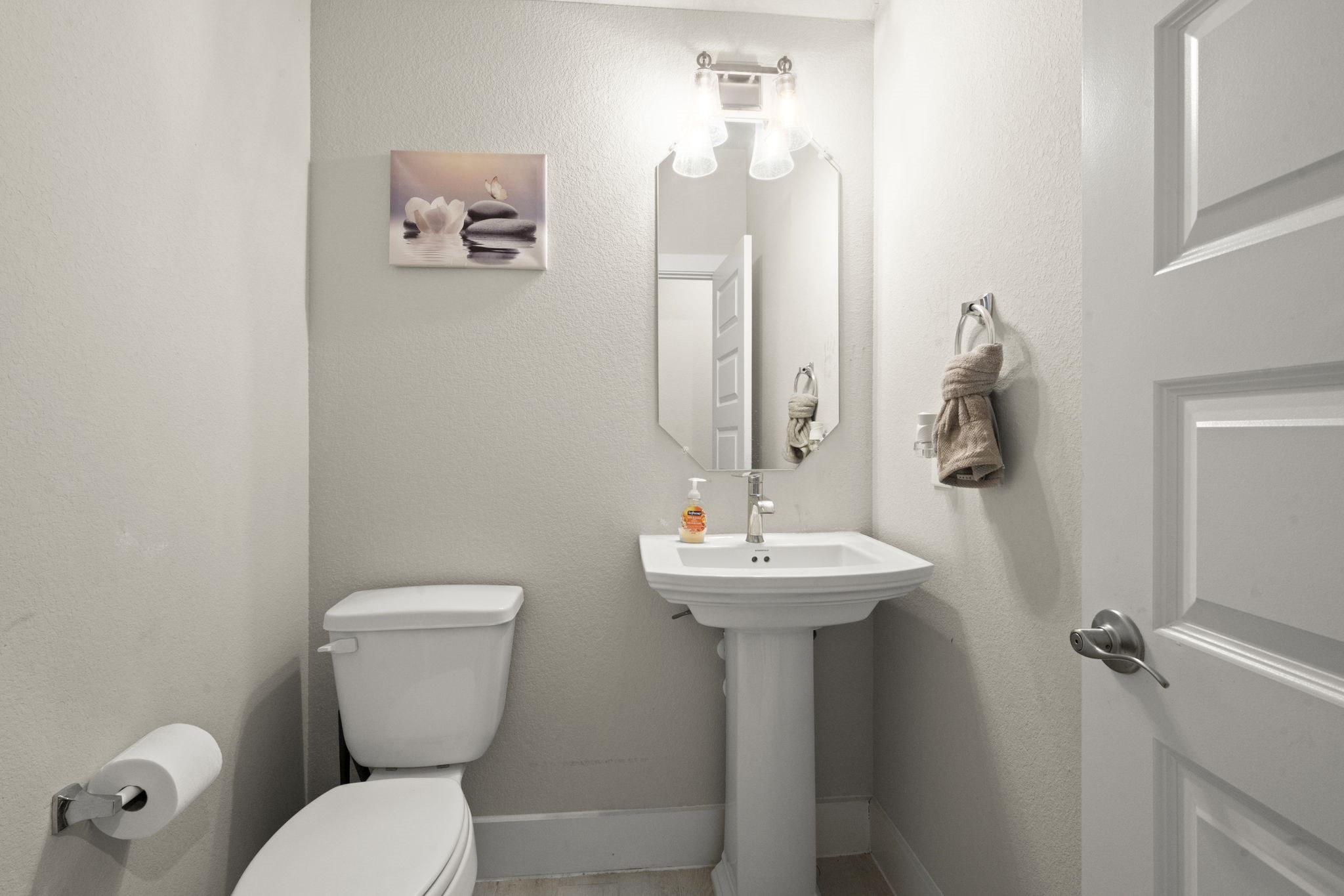 24715 Native Forest Spring, TX 77373 - Photo 9 of 37 This photo shows a compact, modern half-bathroom with a pedestal sink, toilet, and neutral decor. It features a wall-mounted mirror, a simple artwork piece, and a towel ring with a neatly folded towel. The space is well-lit and has a clean, minimalist design.