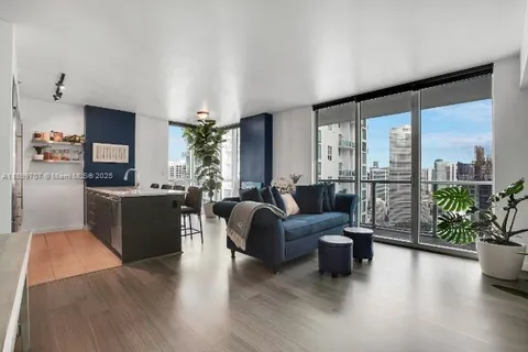 a living room with furniture potted plant floor to ceiling windows and kitchen view