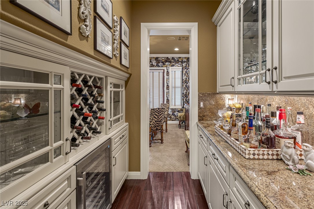 11280 Granite Ridge Drive, Unit 1057 Las Vegas, NV 89135 - Photo 17 of 46 Bar area with wine cooler, light stone countertops, dark wood finished floors, white cabinetry, and glass insert cabinets