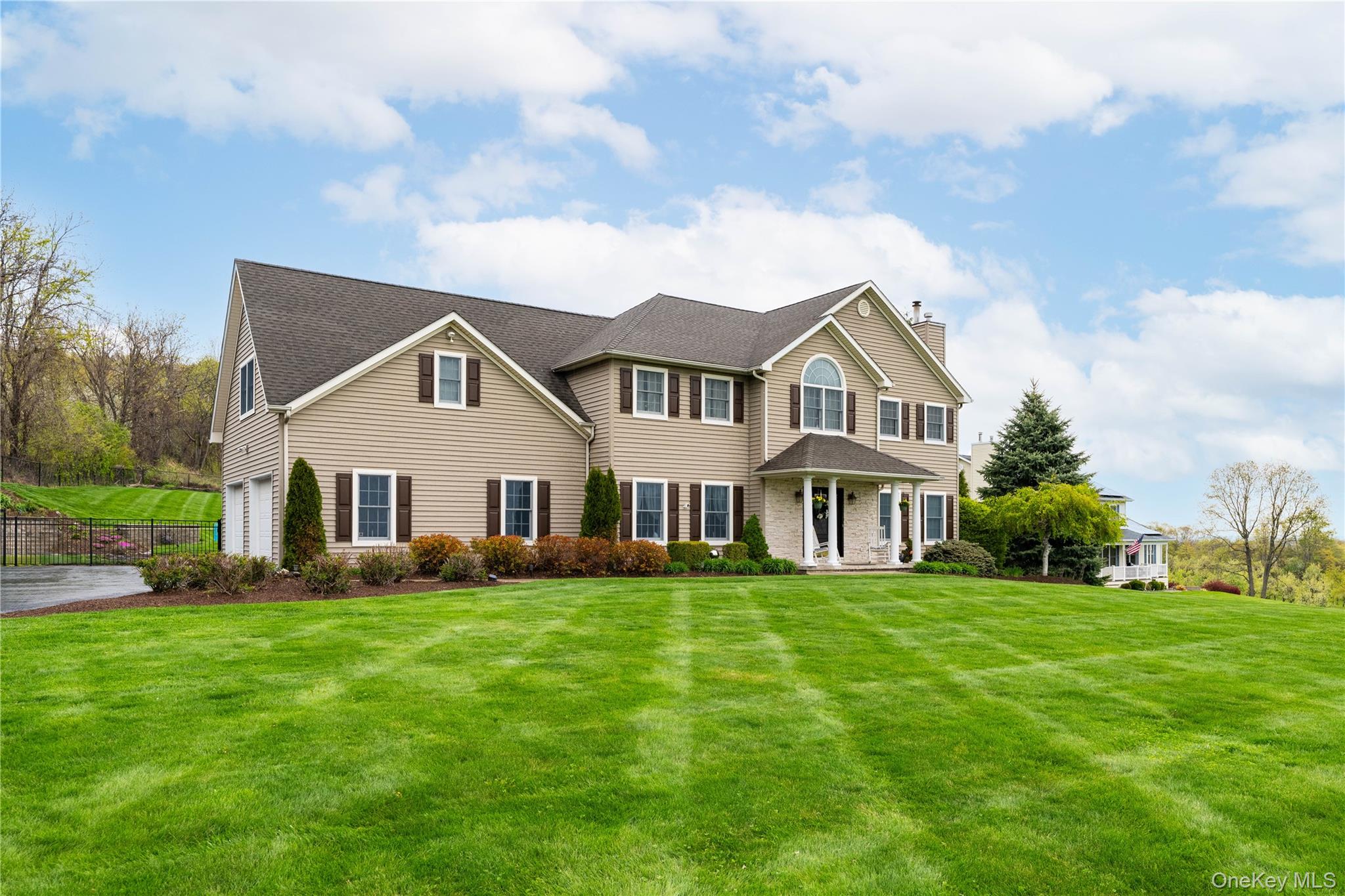 60 Idlewild Road Marlboro, NY 12542 - Photo 2 of 37 Spacious suburban residence featuring a tan siding facade with dark shutters, a built-in multi-car garage, a covered front entry with stone accents, a large manicured lawn, and mature landscaping