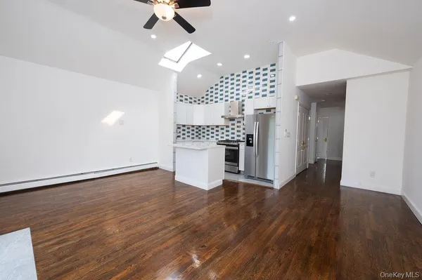 an empty room with wooden floor a ceiling fan a kitchen view and a refrigerator