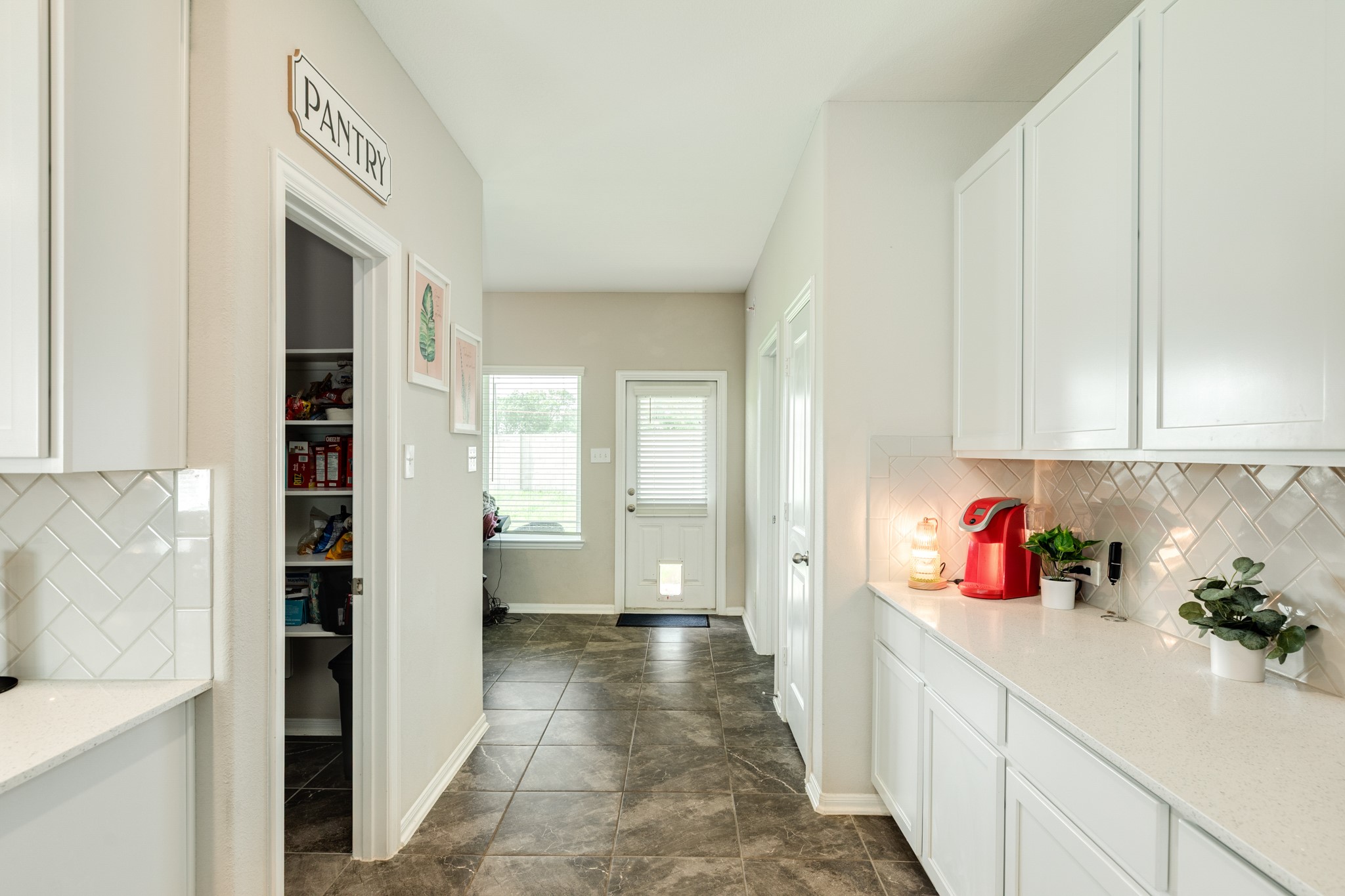 2446 Palisade Crst Drive Rosharon, TX 77583 - Photo 12 of 30 This photo shows a bright kitchen area with white cabinetry, a tiled backsplash, and a visible pantry. It has a clean, modern design with a wood look tiled floor, and leads to a door with natural light coming through. This area can be used for a coffee bar, whiskey bar you decide.