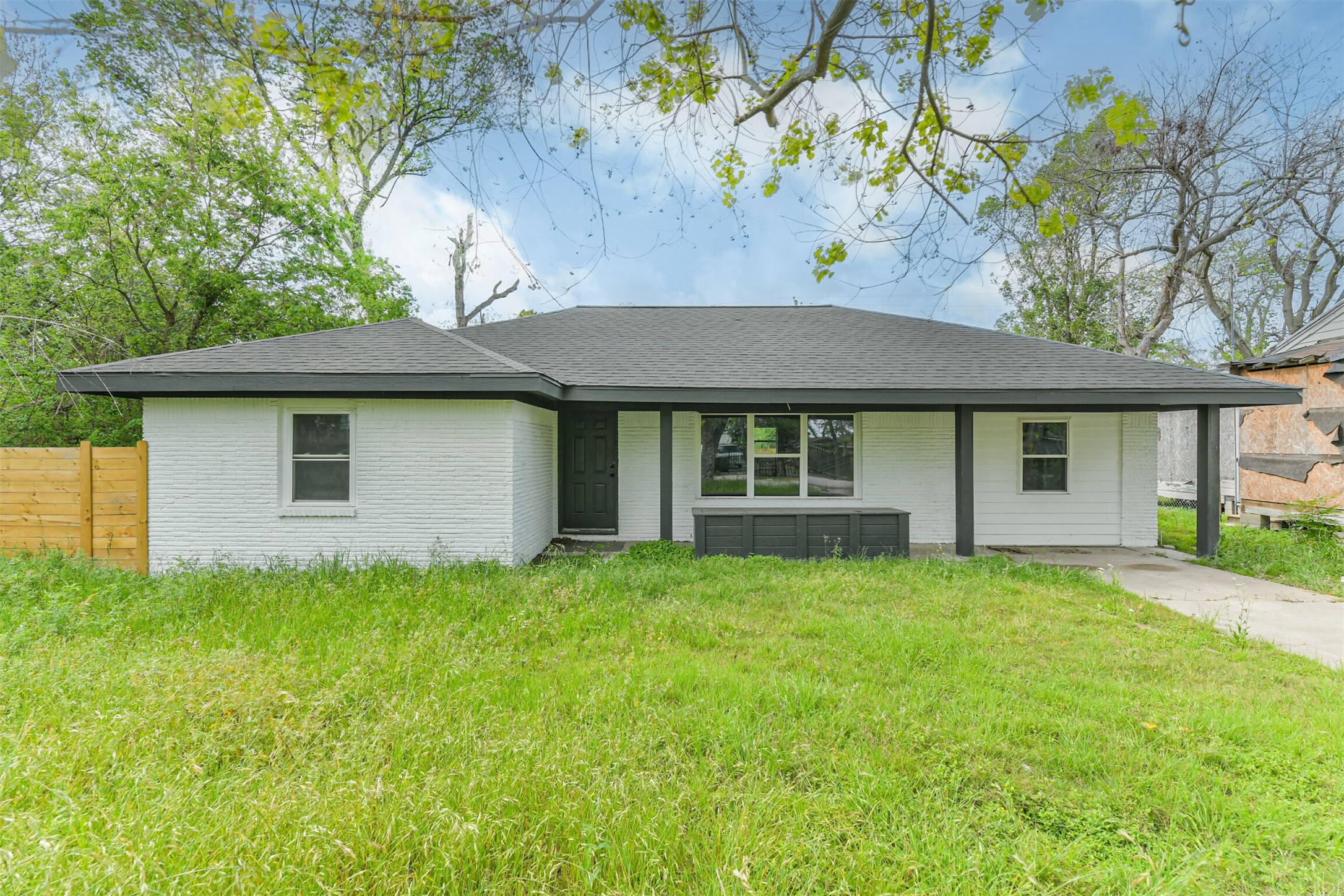 209 Leago Street Houston, TX 77022 - Photo 2 of 24 This is a single-story, white brick home with a dark roof, featuring a covered front porch. It sits on a spacious grassy lot with a wooden fence on one side, surrounded by mature trees for added privacy.