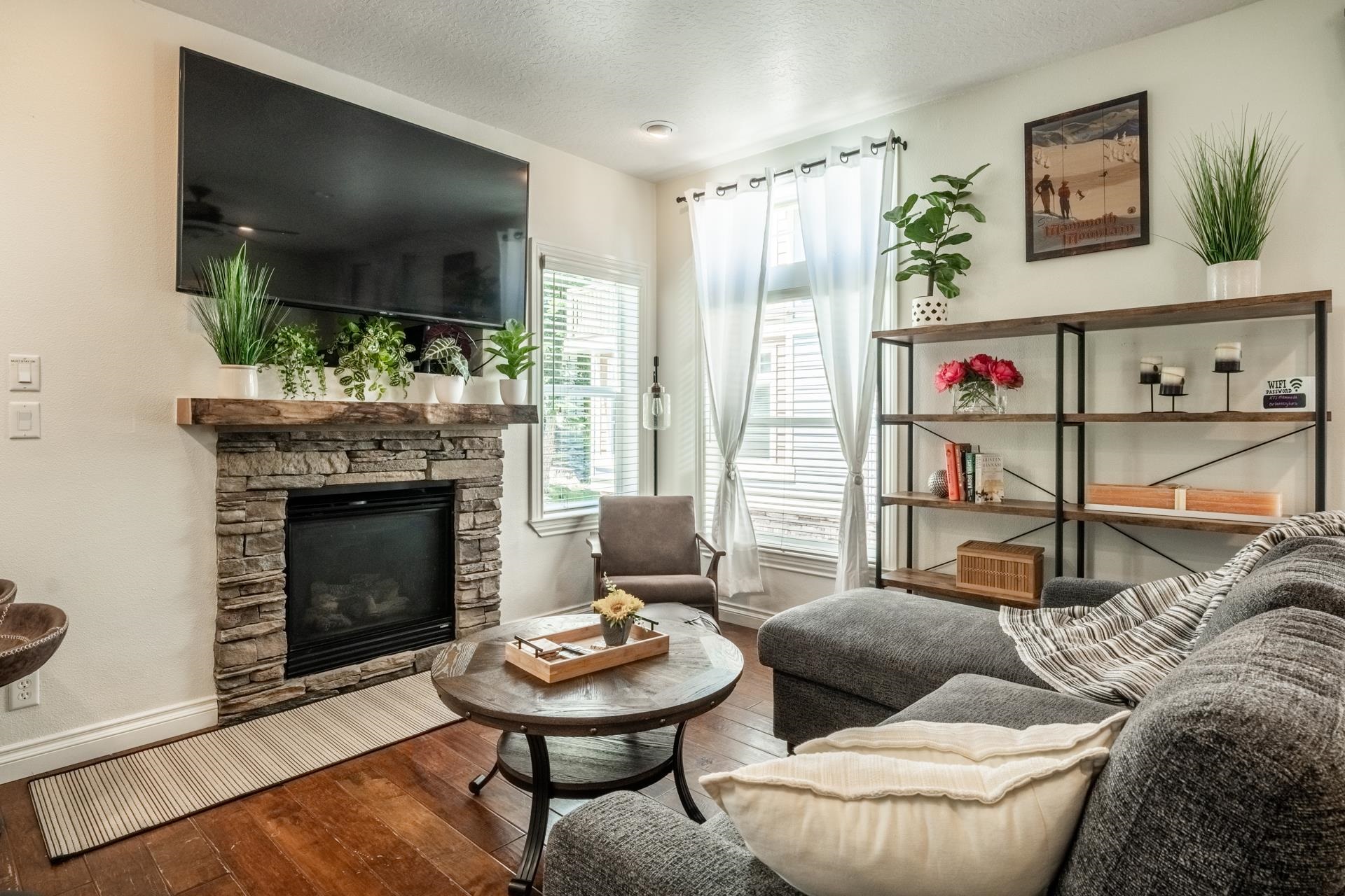 Living area featuring a stone-clad fireplace with a wood mantle, rich wood-finish flooring, and white window blinds