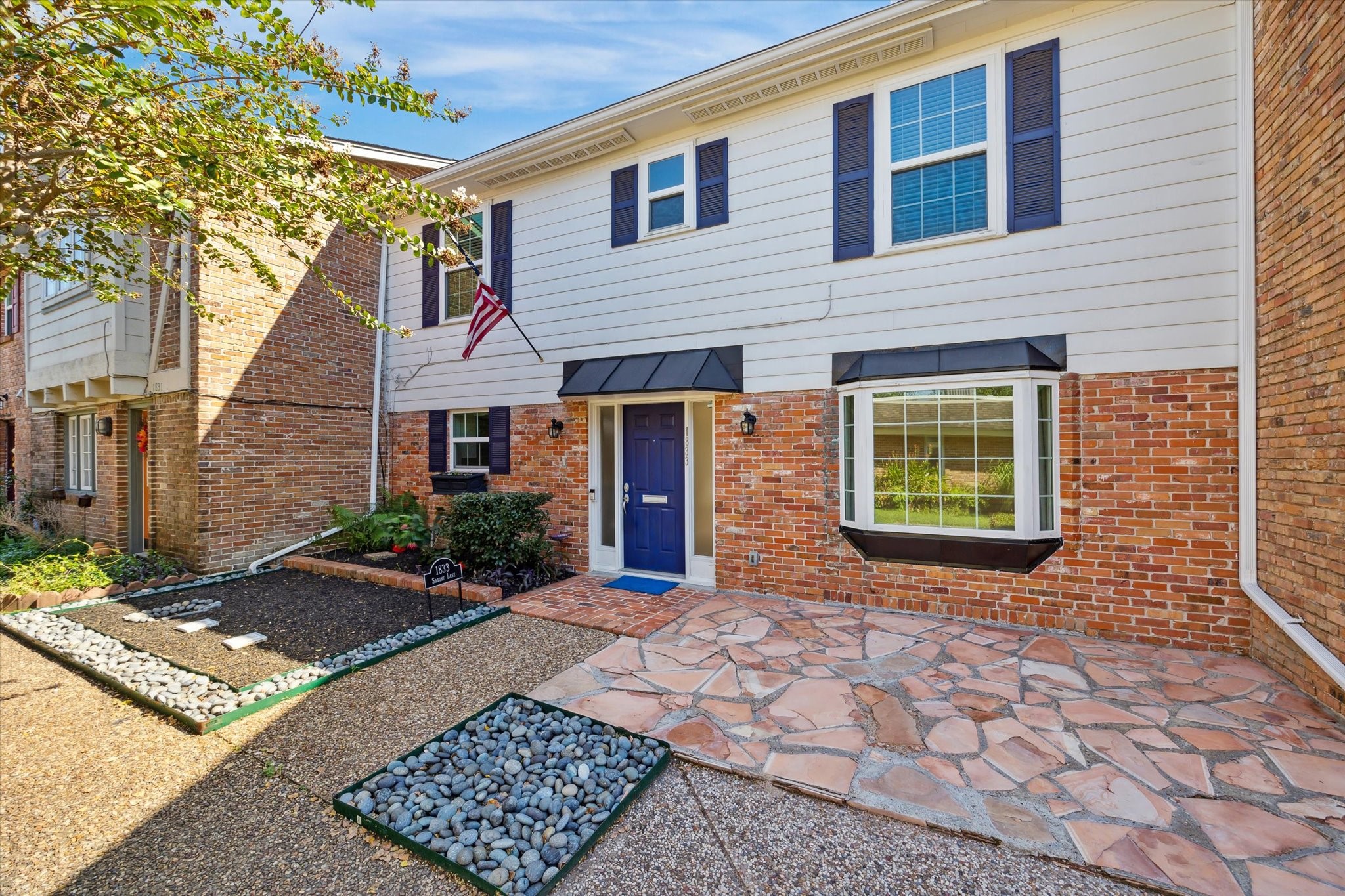 This is a two-story townhome featuring a brick and siding exterior with a welcoming regal blue front door. The entryway includes a neatly landscaped path with stone pavers and pebble accents. In addition to a large bay window on the first floor.