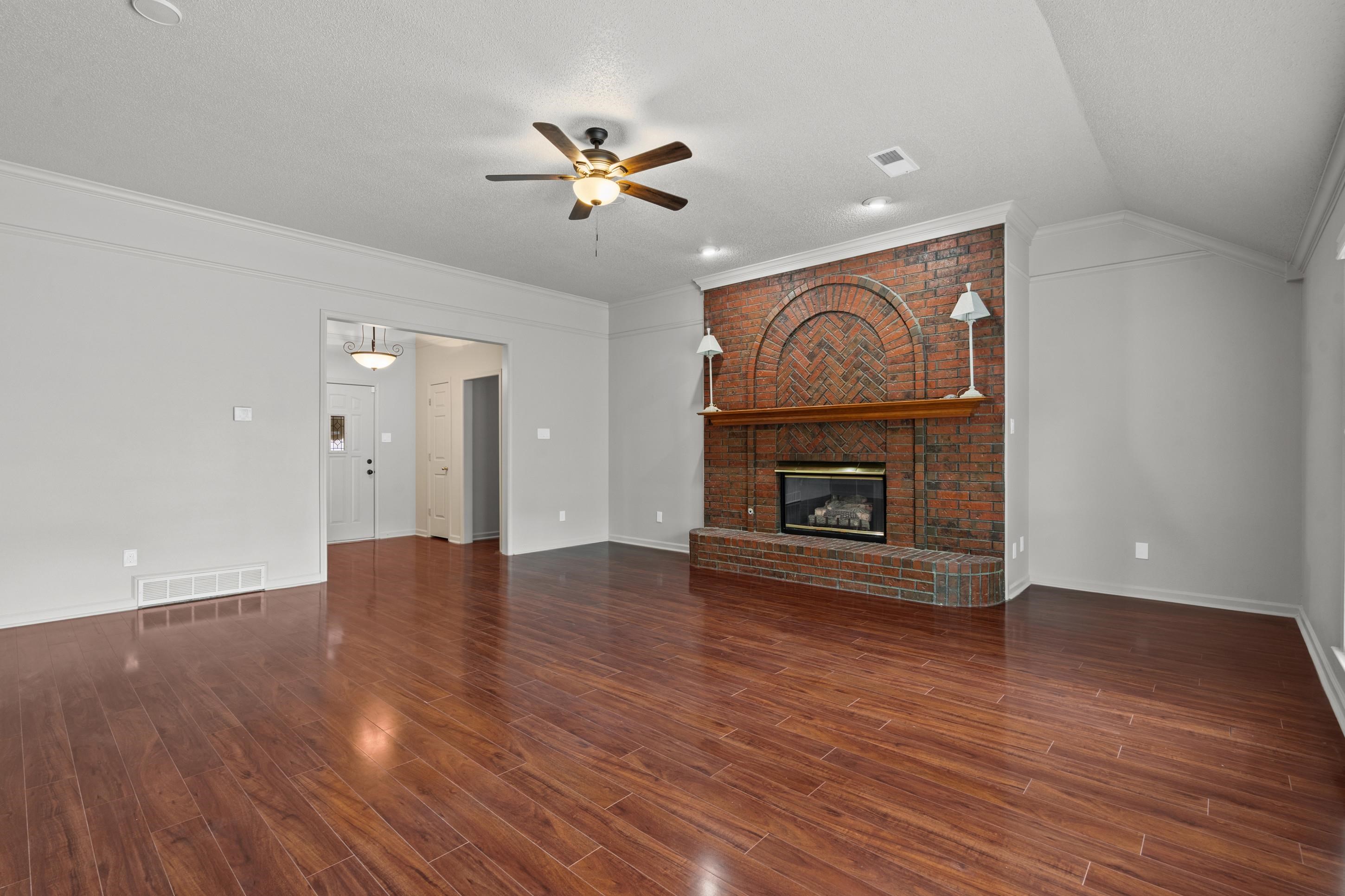 1301 Wolf Lair Drive Collierville, TN 38017 - Photo 10 of 32 Unfurnished living room with ceiling fan, crown molding, a brick fireplace, dark wood-style floors, and vaulted ceiling