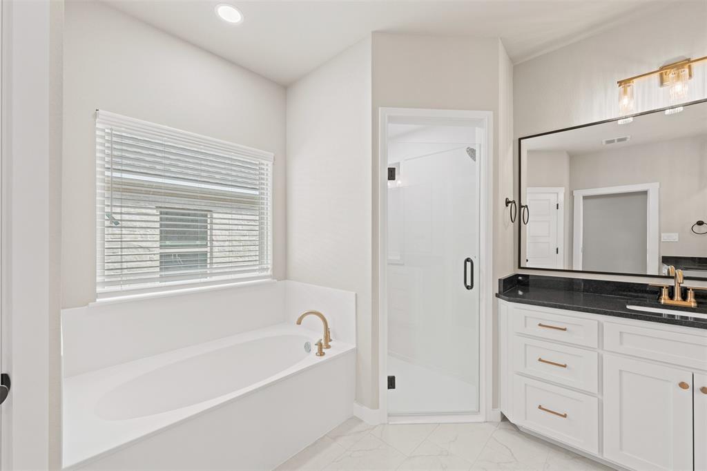 9824 Durango Trail Waco, TX 76712 - Photo 26 of 30 Bathroom featuring a built-in soaking tub with brass fixtures, a walk-in shower with a frameless glass door, a double vanity with a dark countertop and gold-toned hardware, and a large wall-mounted mirror