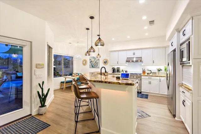 a kitchen with stainless steel appliances kitchen island granite countertop a refrigerator a stove a dining table and chairs with wooden floor