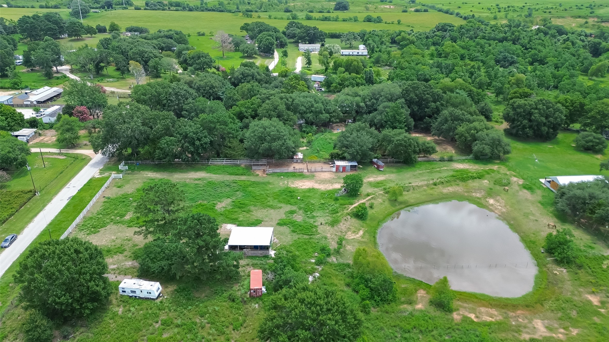 35535 Windmill Road Hempstead, TX 77445 - Photo 37 of 41 This aerial photo showcases a spacious rural property with lush greenery, a sizable pond, and several structures, including a small house and a trailer. The area is surrounded by trees and open fields, offering a peaceful, countryside setting with nearby road access.