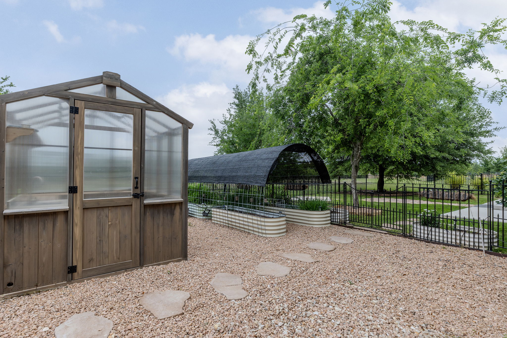 128 Strada Luca Florence, TX 76527 - Photo 34 of 40 Dedicated garden area designed for functionality and growth, featuring a wood-frame greenhouse with translucent panels, gravel pathways with stepping stones, raised metal garden beds, and a covered shade structure, all enclosed for an organized and productive outdoor space.