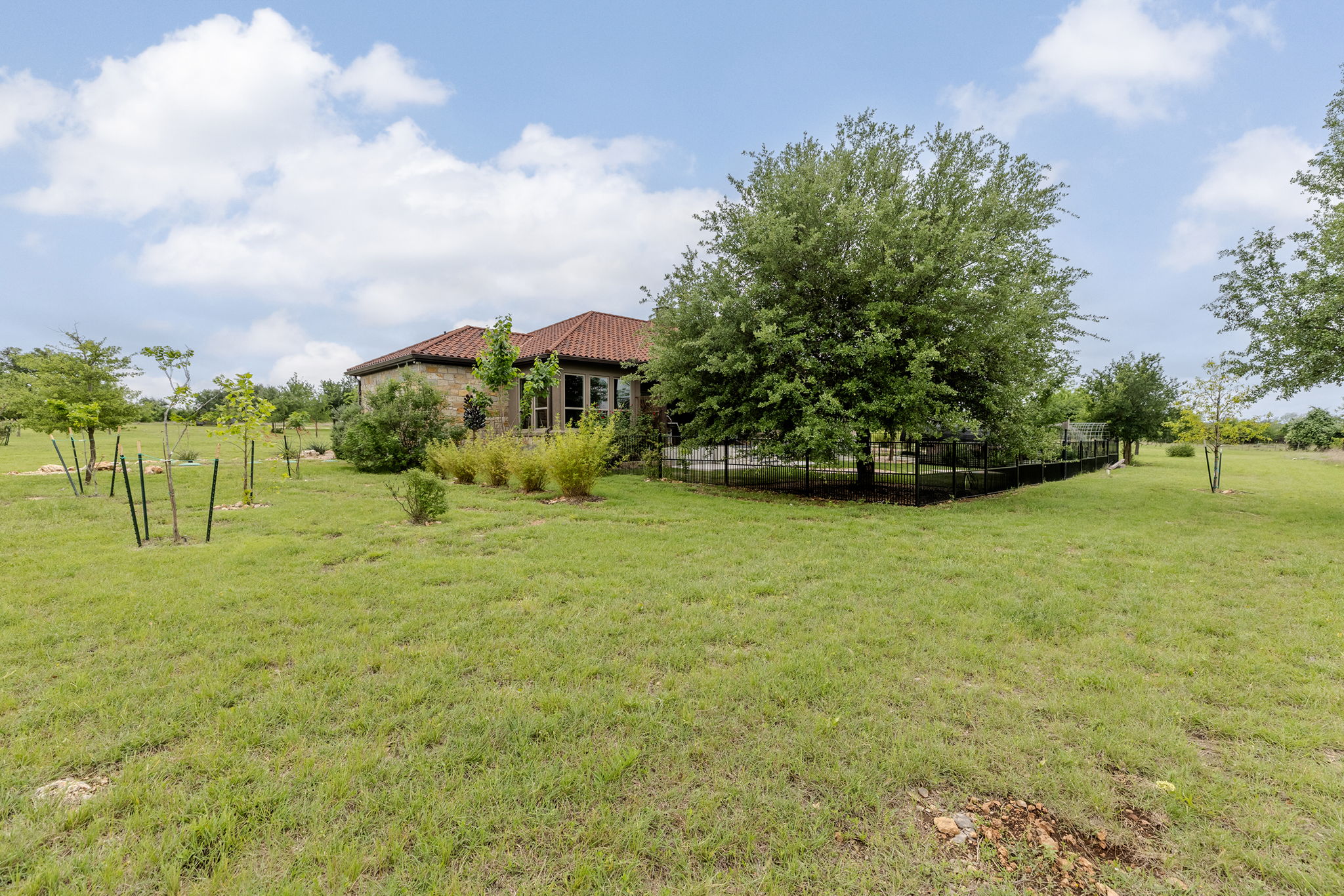 128 Strada Luca Florence, TX 76527 - Photo 35 of 40 Expansive, park-like yard surrounding a stone-clad residence with a terracotta-style tile roof, complemented by mature trees and a black perimeter fence, offering wide-open space, privacy, and scenic natural views.
