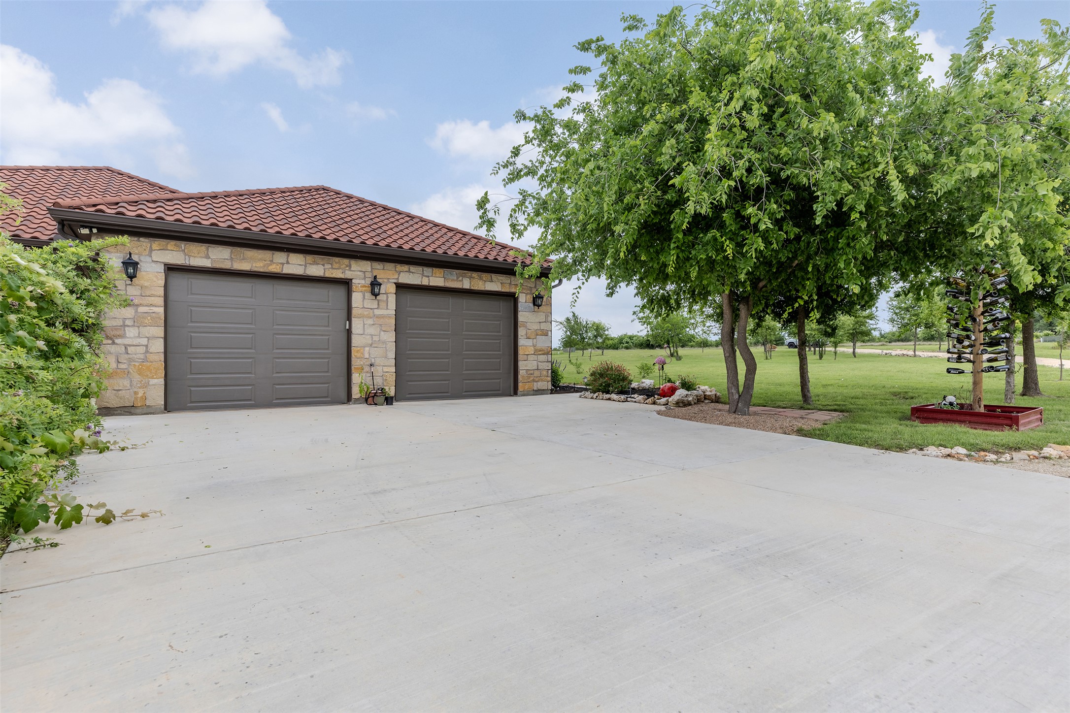 128 Strada Luca Florence, TX 76527 - Photo 4 of 40 Stone facade garage with dual doors and a terracotta-style tile roof, complemented by a spacious concrete driveway and mature trees providing shade and privacy.
