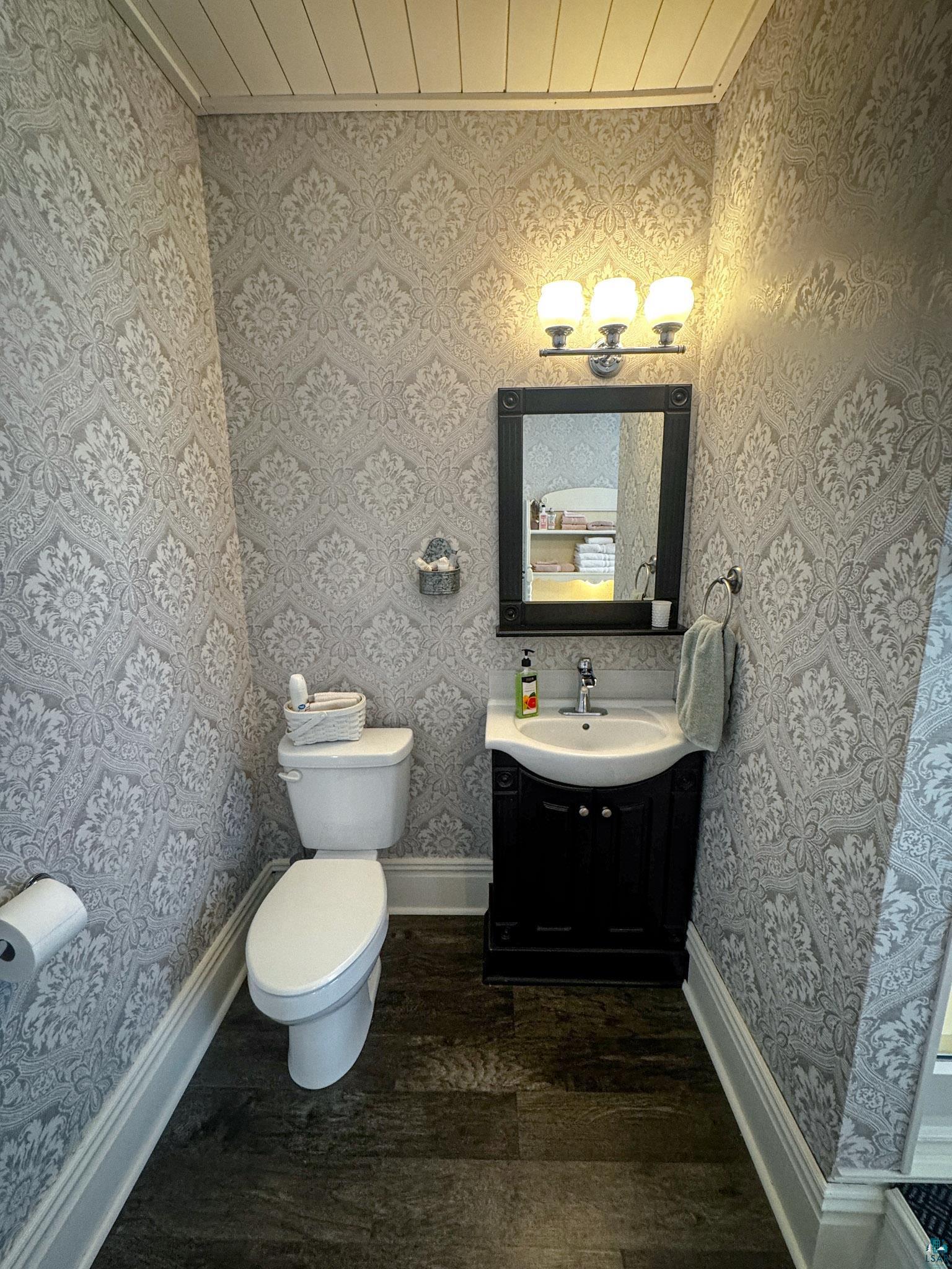303 East 5th Street Washburn, WI 54891 - Photo 25 of 34 Powder room featuring decorative wallpaper, wood-finish flooring, a contemporary vanity with an integrated sink, a dark framed mirror, and a three-light fixture