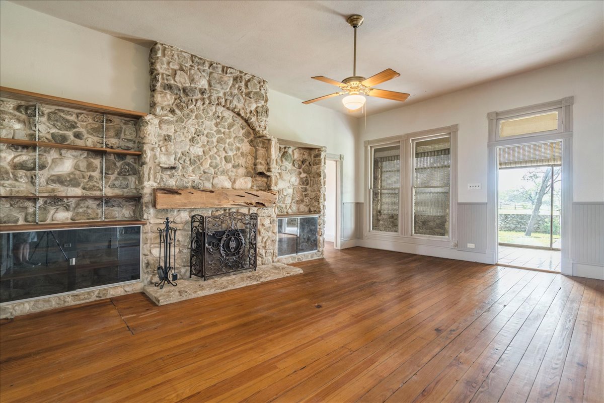 6400 Cypress Road Kyle, TX 78640 - Photo 13 of 33 Spacious room featuring a stone fireplace with a substantial timber mantel, built-in shelving, wood-finish flooring, multiple windows, and a ceiling fan